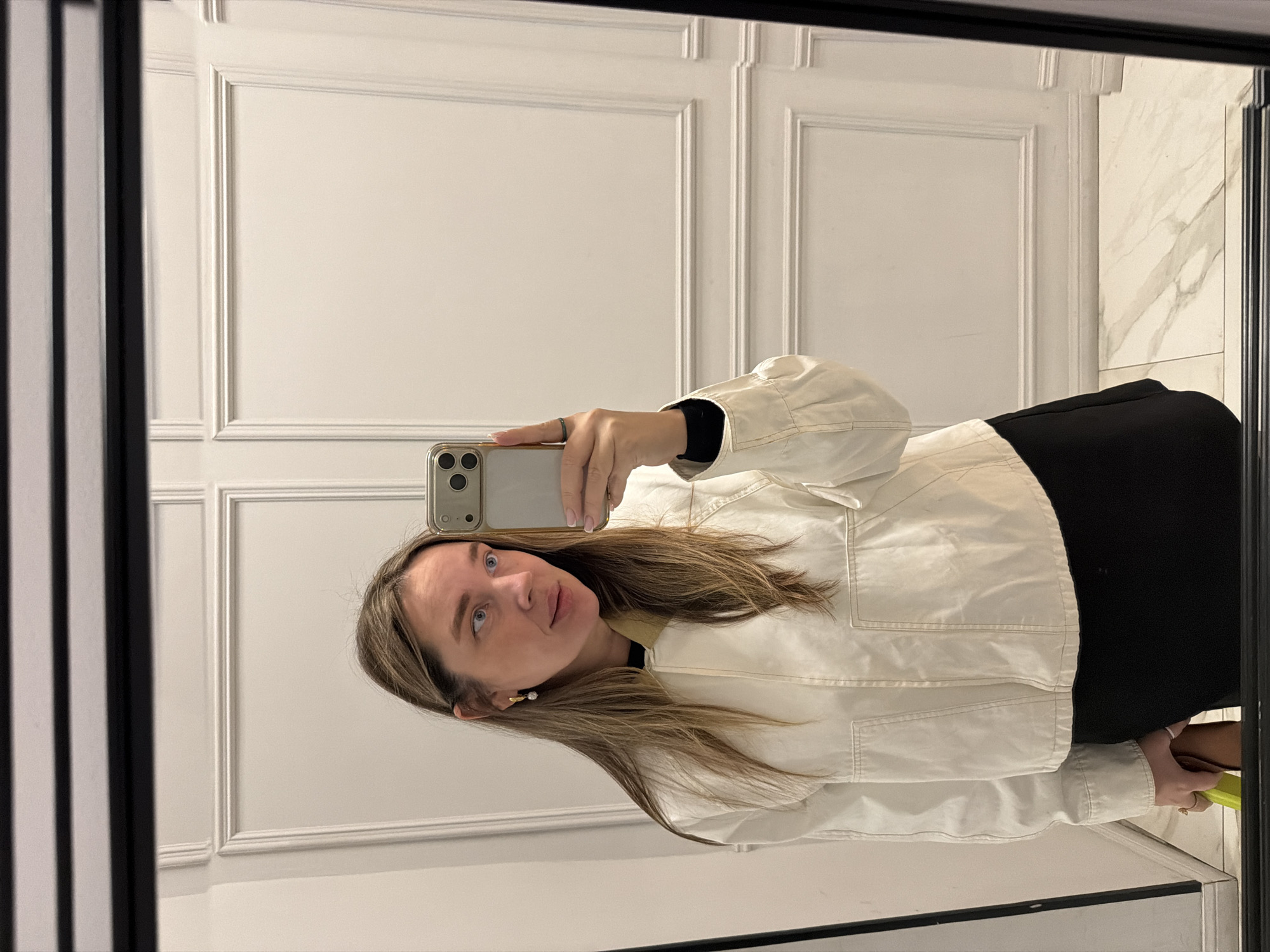 A woman is taking a selfie in a mirror. The mirror frame is black. The wall behind the woman and mirror has a lower portion with light marble tile. Above the marble, the wall is white with raised rectangular trim pieces. The woman is wearing a black top, black skirt and a white jacket. She is holding a smartphone in her right hand with gold colored sides and three camera lenses. She has medium length brown hair and is wearing an earring on her left ear. Based on the style of the decor, this is likely a public space in Moscow, Russia.