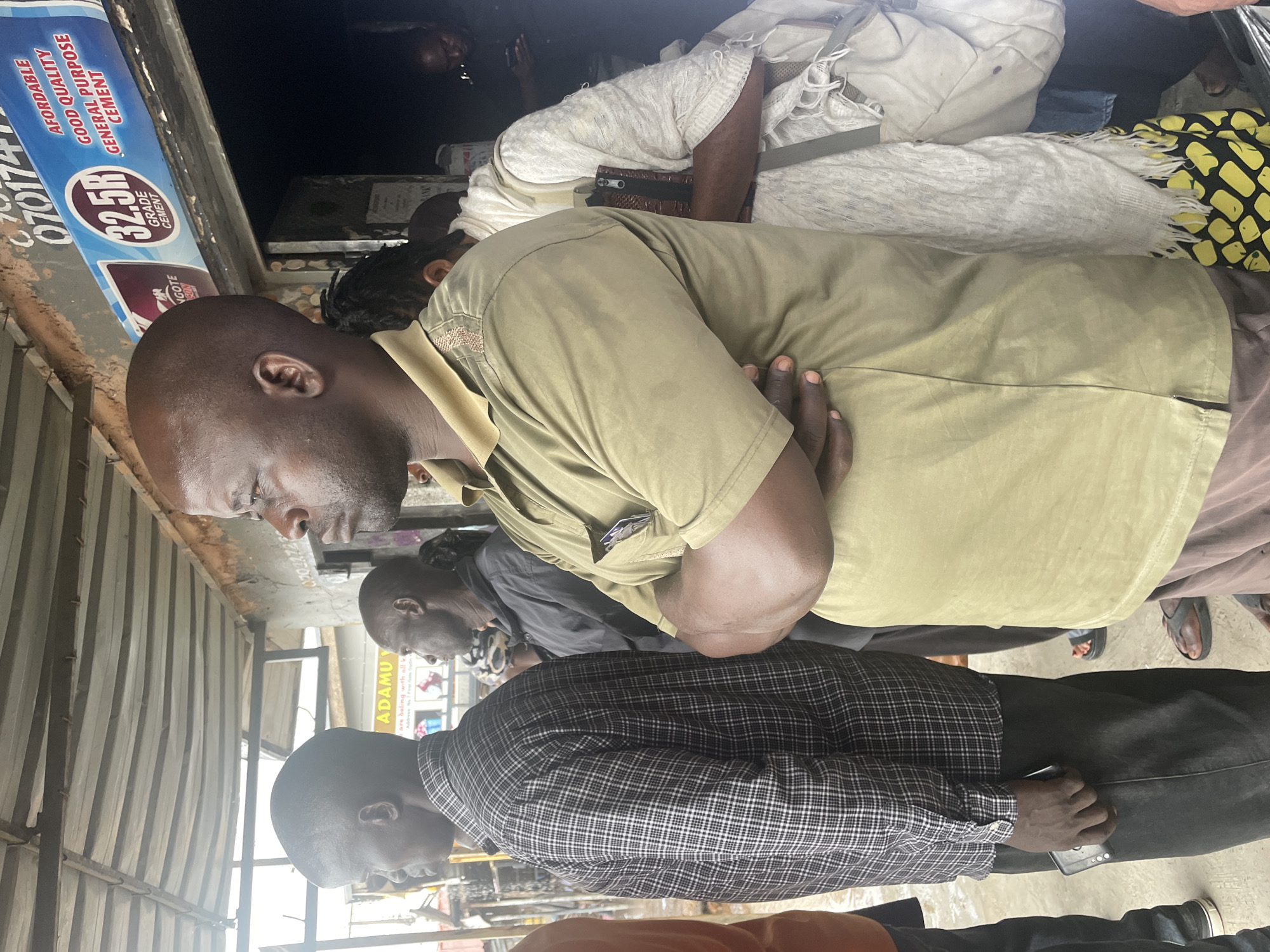 The image captures a street scene in what is identified through the text as the city of Gura Top, Nigeria. A group of at least four African men are present. One man is prominently dressed in an olive green shirt and brown pants, with his arms crossed. Below him is another man in a plaid shirt and dark pants, holding a mobile phone. Slightly behind the man in green is another man wearing a gray shirt. Parts of additional people can be seen at the bottom and top of the frame. In the background, a sign advertises "32.5R GRADE CEMENT" with text mentioning "AFFORDABLE," "GOOD QUALITY," and "GENERAL PURPOSE CEMENT." Another sign appears to say "ADAMU" with the additional text "are beling with all k." The location is along a street lined with corrugated metal storefronts.