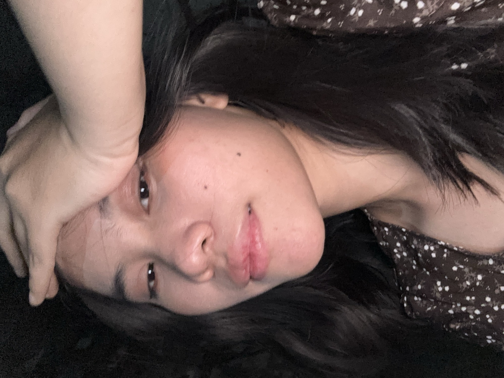A young woman with dark hair is seen in a close-up shot. She is wearing a dark brown, short-sleeved dress with a white floral pattern. Her right hand is placed on her forehead, and her left arm is visible across her face. The background is dark, indicating a low-light environment. The image appears to be a selfie taken indoors. The context of East Jakarta, Indonesia, is not visually present in the image.