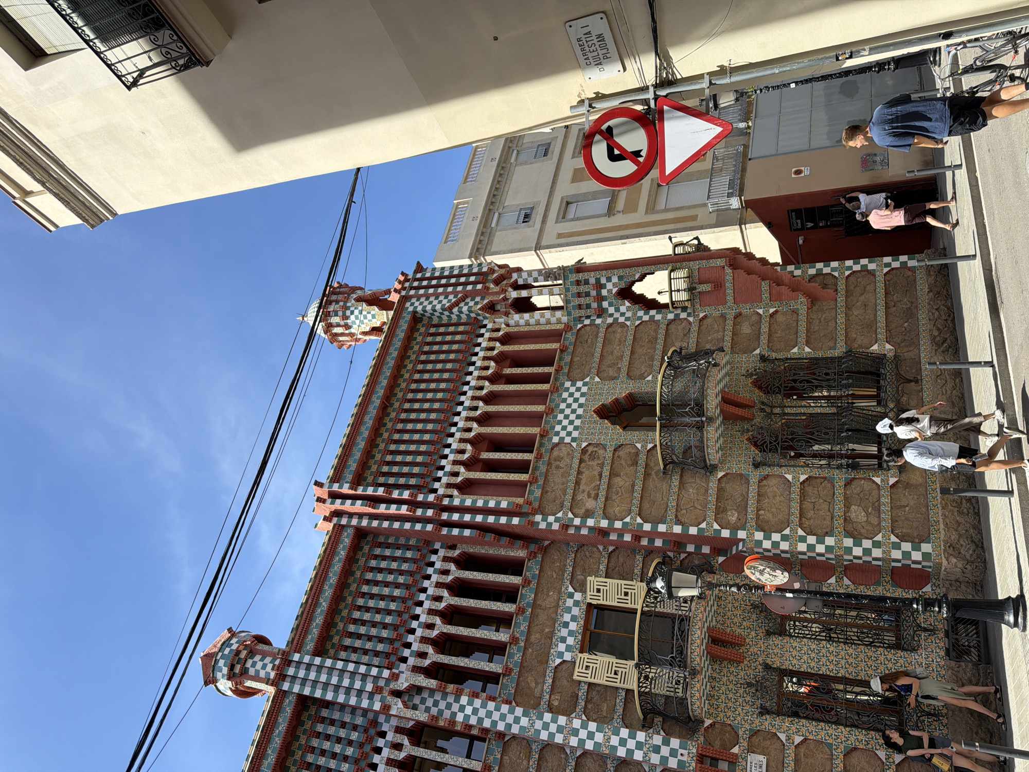 In the city of Barcelona, Spain, a colorful, intricately tiled building with red, green, and white checkerboard patterns stands under a bright blue sky. The building's facade features arched windows, decorative balconies with wrought-iron railings, and a unique tower with a checkered dome. To the right, a lighter-colored building casts a shadow on the street. A street sign reads "CARRER D'AULESTIA I PIJOAN." Several people are walking on the sidewalk in front of the building, including a group of young women on the left and individuals further down the street. Traffic signs, including a "no left turn" symbol and a yield sign, are visible on a pole. A black lamppost with a mirror is also present on the sidewalk. The scene captures a vibrant urban streetscape on a sunny day.