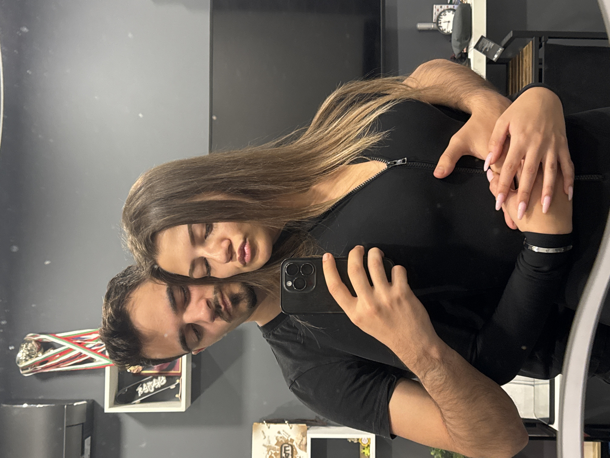 This intimate image is a mirror selfie featuring two young adults, a man and a woman, captured indoors. The photo shows the couple in a close embrace, their heads together, with both individuals having their eyes closed or nearly closed, conveying a sense of peace and affection.
The woman has long, straight brown hair with lighter highlights and is wearing a black zippered top. Her left hand, adorned with long, light pink pointed nails, rests on the man's arm, and a silver bracelet, possibly a Cartier Love bracelet, is visible on her wrist. The man has dark hair, a beard or stubble, and is wearing a black T-shirt. His right hand holds a black smartphone (likely an iPhone) to take the picture, reflecting their image in the mirror. His arms are noticeably hairy.
The background, seen as a reflection, reveals a domestic setting with a dark gray wall. A large black screen or television is mounted on the wall. To the left, a white shelf holds a small silver trophy decorated with ribbons in white, green, and red, which are the colors of the Bulgarian flag, consistent with the location context of Sofia, Bulgaria. Below the trophy, another shelf displays a miniature skateboard with the word "NAZAR" written in white. Further back, a wall clock and other indistinct objects are visible. The image captures a serene and affectionate moment between the two subjects.