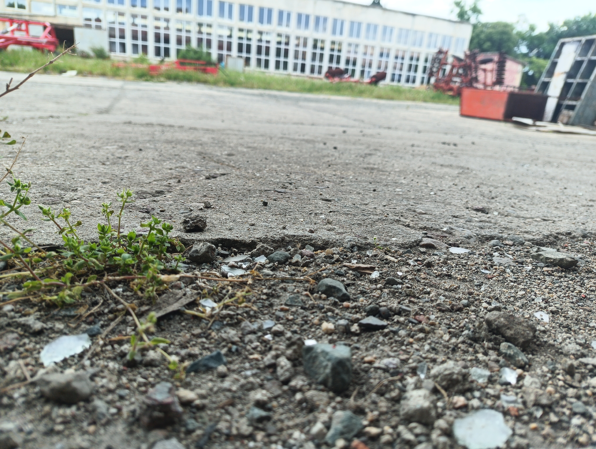 The image shows a close-up view of a cracked concrete and gravel surface in an outdoor environment. Small green plants sprout from the edge of the concrete. Scattered rocks and shards of glass are visible among the gravel. In the background, a long, white building with multiple windows is partially obscured by machinery and other indistinct objects. The scene appears to be located in an industrial or neglected area of an unknown city. There are no people visible.