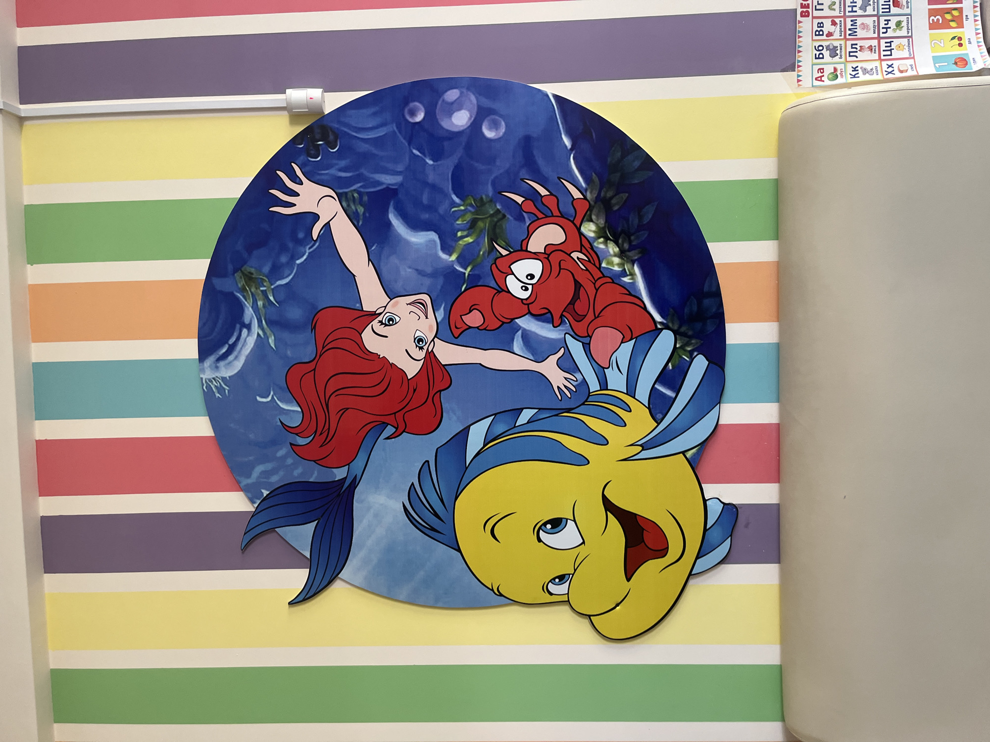 This image depicts a colorful wall decoration featuring characters from Disney's "The Little Mermaid". Ariel, Sebastian, and Flounder are prominently displayed against a blue oceanic background, cut out in a round shape. The wall itself is painted with horizontal stripes of various colors, including yellow, green, orange, purple, and pink. A portion of a white chair or couch is visible on the right side of the frame. The upper right corner shows an educational chart with Russian letters and numbers. The location of this image could be in the city of Ulyanovsk, Russia.
