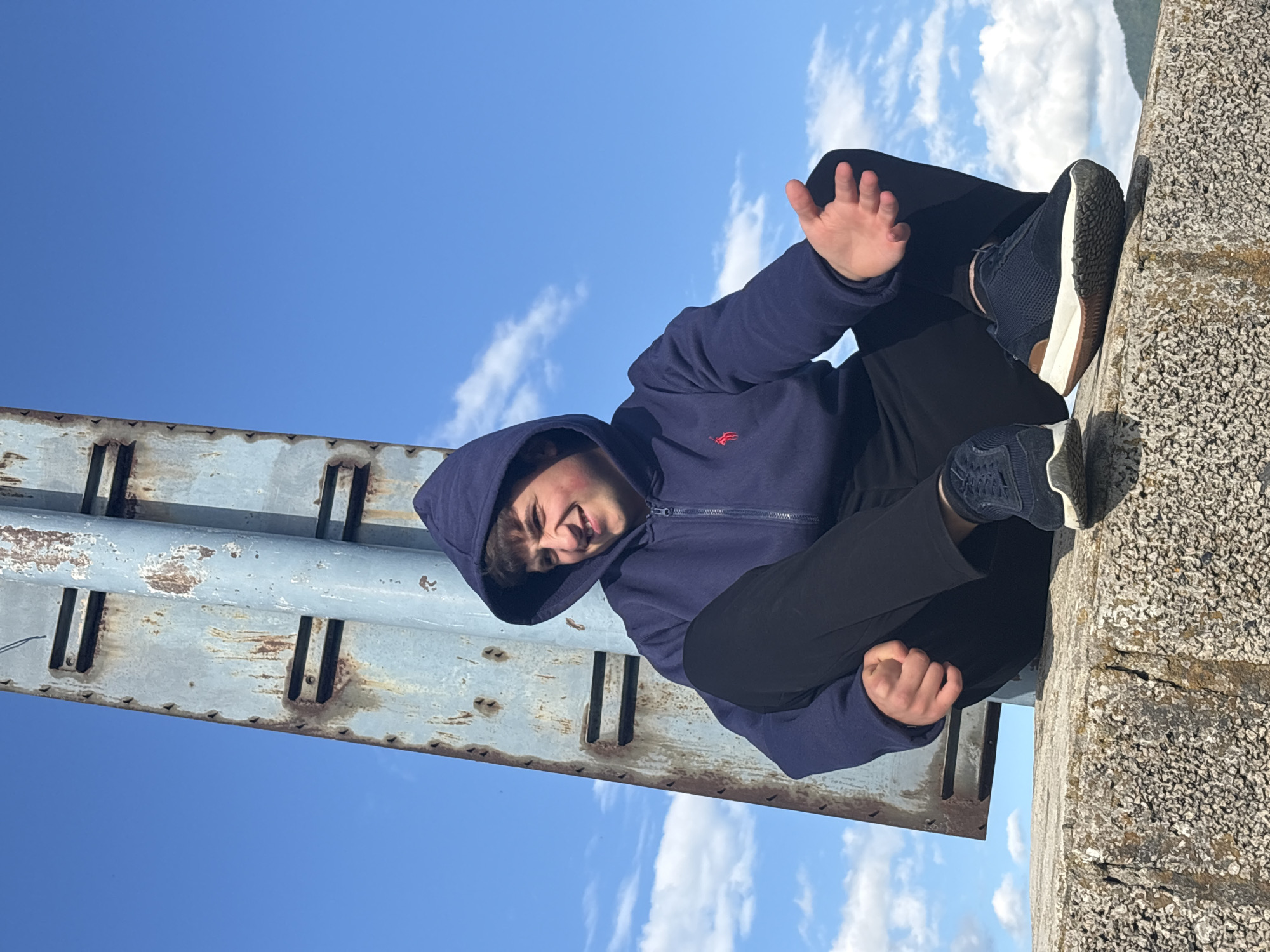 A person is sitting on a concrete structure next to a rusted metal beam in Alaverdi, Armenia. The person wears a navy blue hooded sweatshirt with a red logo, black pants, and navy blue sneakers with white soles. The person's right hand is raised. The background features a blue sky with scattered white clouds. The person is resting their feet on a rough concrete surface.
