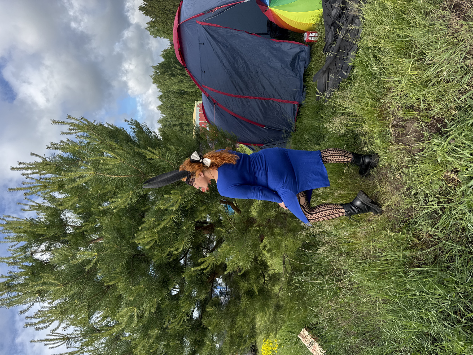 A woman with long, curly red hair, adorned with a white bow and black horn-like headwear, kneels on grassy ground in an outdoor setting in Большая Кивара, Russia. She wears a long-sleeved royal blue dress, black fishnet stockings, and black combat boots. A large evergreen tree is prominent to her left. In the background, a blue and red dome tent is visible, next to a colorful, possibly rainbow-striped umbrella or shelter, and a small red metallic container. The sky is partially cloudy with patches of blue. The scene suggests a daytime outdoor event or camping trip, with the woman posing for a photograph. Faint, illegible text is visible on a piece of wood in the lower right foreground, appearing to start with "Яго".
