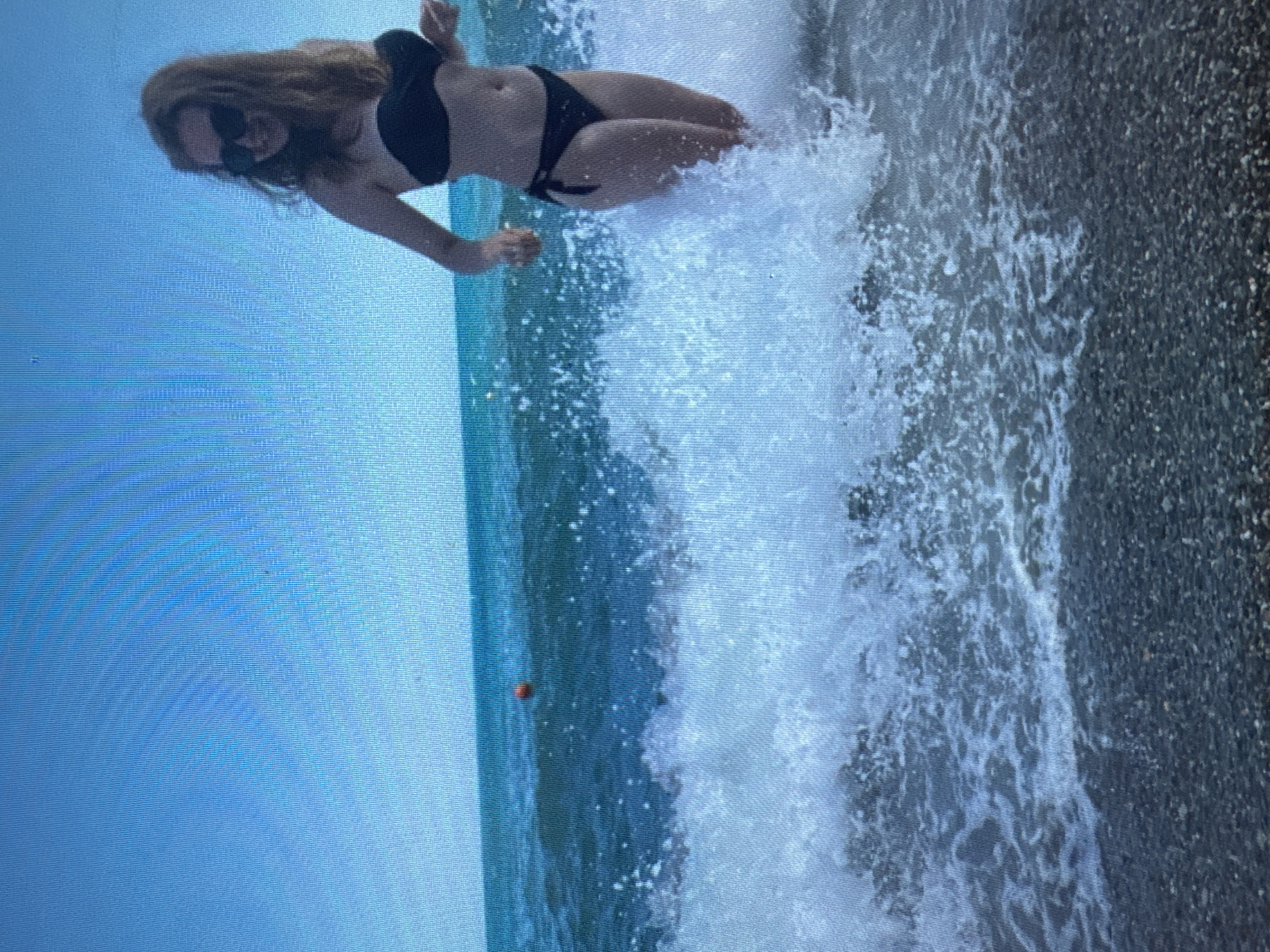 A woman with long, reddish-blonde hair and sunglasses, wearing a black bikini, stands in the ocean amidst breaking waves. She is partially submerged in the water, with a wave cresting around her waist and chest. The background consists of a clear blue sky and the teal-colored ocean. A small, orange buoy is visible in the distance on the water. The foreground shows a pebbled beach with the waves lapping at the shore.