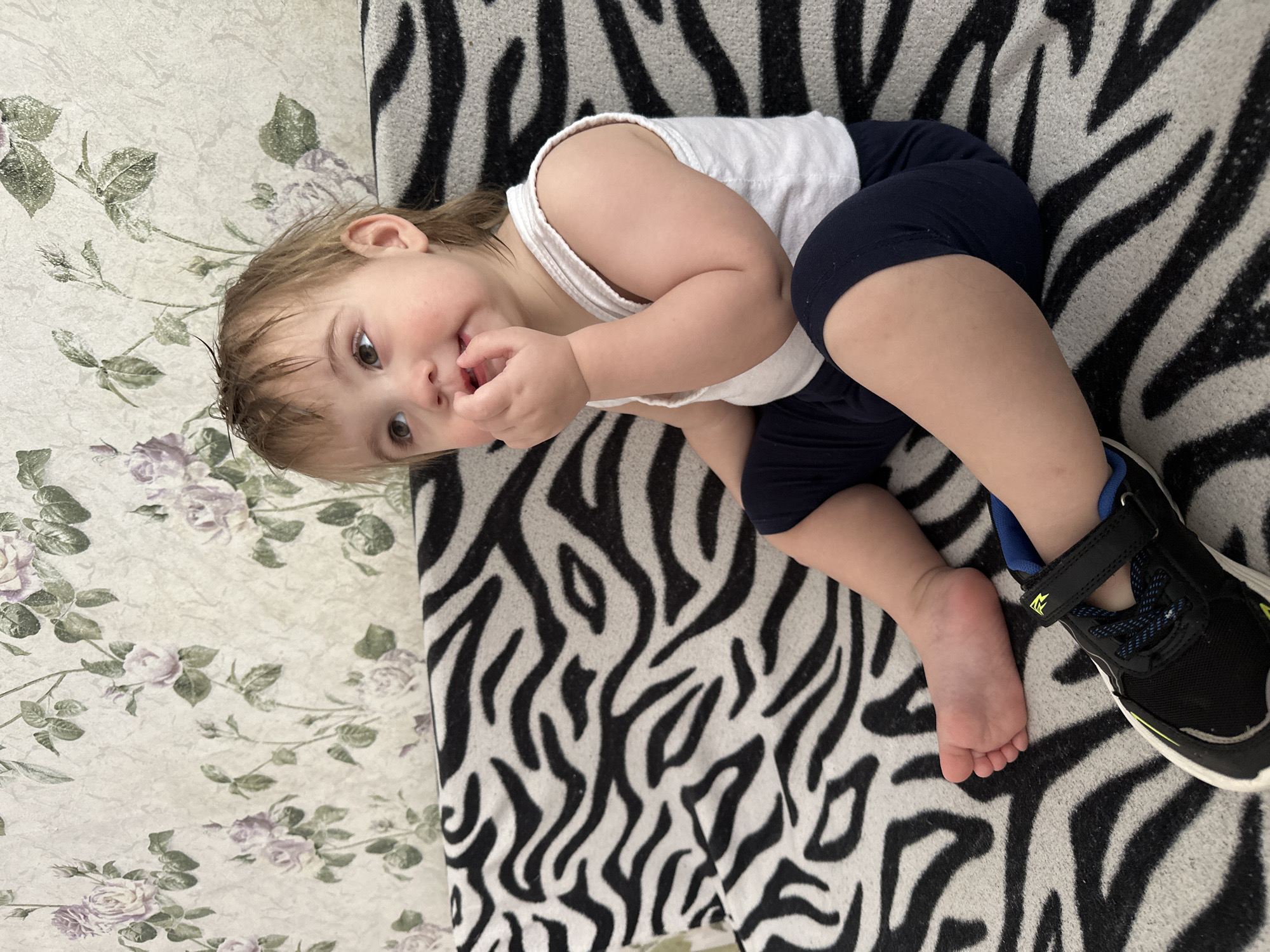 A toddler is depicted sitting upright indoors. The child has light brown hair, appearing somewhat damp or disheveled, and brown eyes directed upwards and to the viewer's left. The child's right hand is at the mouth, with fingers visible. The child wears a white ribbed sleeveless top and dark blue shorts. The left foot is bare, while the right foot is fitted with a black athletic shoe featuring blue accents and a yellow stripe. The child is positioned on a surface covered with a black and white zebra-print blanket or upholstery. Behind the child, a wall is visible, covered in wallpaper patterned with light purple roses, green leaves, and stems on a light, textured background. The precise location within the city of Chaikovsky, Russia, cannot be determined from the visual evidence presented in the image.