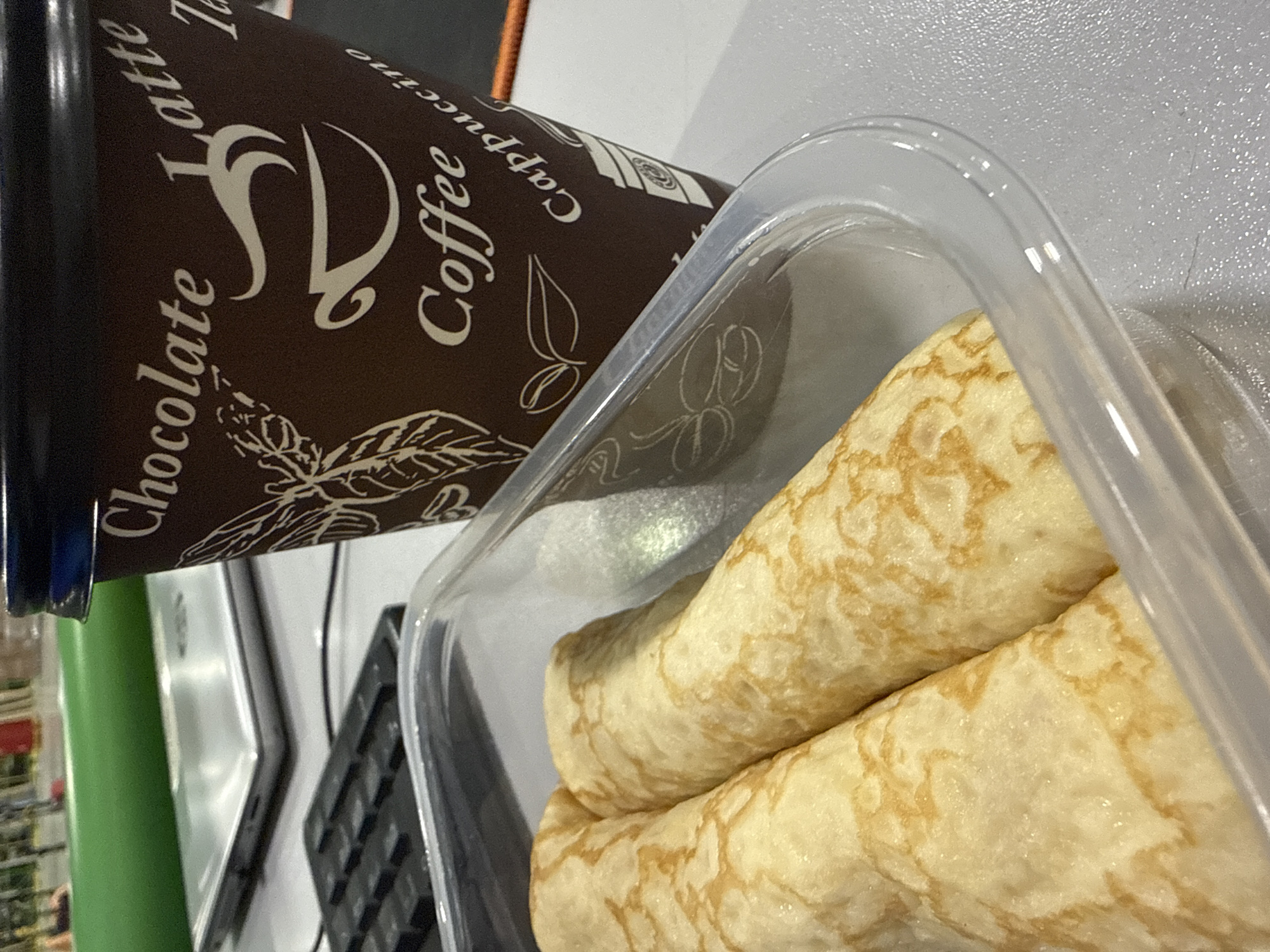 This image, taken in Lupanovo, Russia, captures a close-up of a desk setup featuring food and drink. The main subjects are a dark brown disposable coffee cup and a clear plastic container holding two rolled crepes or pancakes. The coffee cup features white text including "Chocolate," "Latte," "Coffee," "Cappuccino," and "Tea," alongside a stylized logo and leaf illustrations. The crepes, light golden-brown with darker marbled patterns, are rolled and presented inside the transparent container. In the background, elements of a workspace are visible, including a section of a black keyboard and a reflective silver object, possibly a laptop or tablet. A bright green cylindrical item is partially obscured on the left side of the frame. All items rest on a light-colored, textured surface, likely a desk or countertop, suggesting an indoor setting for a meal or snack break.