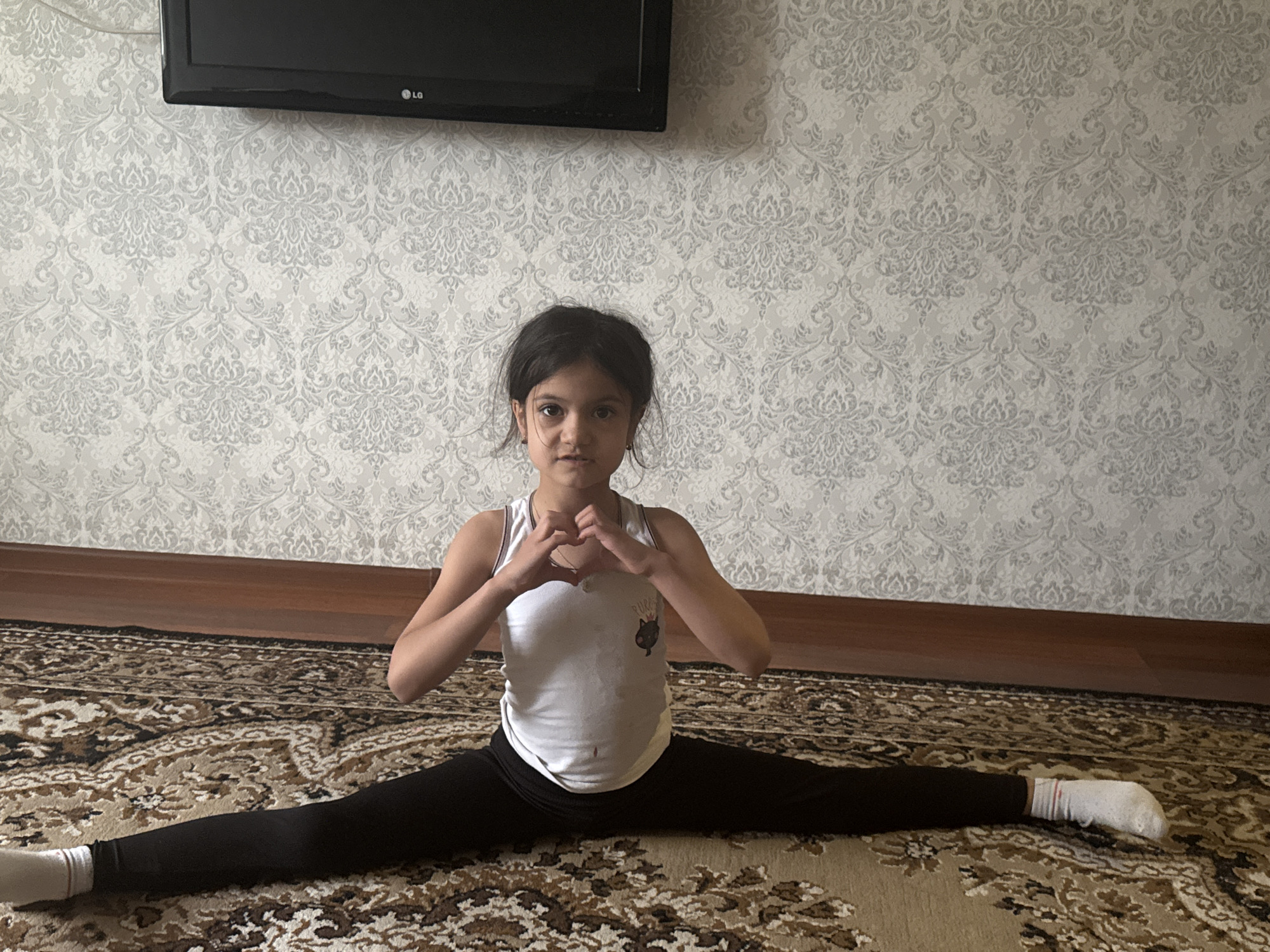 A young girl is featured performing a full splits stretch on a patterned rug indoors. She is positioned centrally in the frame, looking directly at the camera with a neutral expression. Her hands are brought together in front of her chest to form a heart shape. She is dressed in a white sleeveless top, black leggings, and white socks.
The background consists of a wall covered with light-colored wallpaper featuring an intricate gray damask or floral pattern. A black flat-screen television, displaying the "LG" logo, is mounted high on this wall. Below the wallpaper, a dark wooden baseboard separates the wall from the floor. The floor is covered by a large rug with a complex, ornate pattern in shades of brown, beige, and black.
The scene is brightly lit, suggesting daytime. The setting appears to be a home environment, possibly a living room or bedroom. No specific weather conditions are discernible from this indoor view. The girl's posture suggests an activity related to gymnastics or flexibility training. A small graphic, possibly depicting a cat's head and partially visible text that seems to include "P.U.R.R", is on her white top.