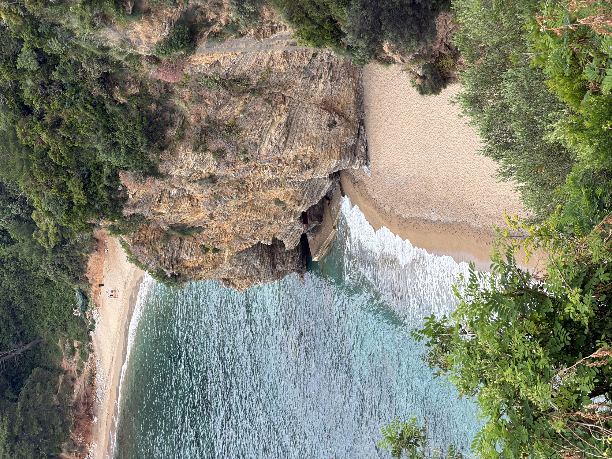 This high-angle, aerial perspective image captures a picturesque, secluded cove on the coast of Budva, Montenegro. The scene is dominated by a clear, calm sea of varying shades of turquoise and blue, meeting two sandy beaches that flank a prominent rocky headland.
The left side of the image reveals a long, crescent-shaped sandy beach bordered by lush green foliage and a dirt track. On this beach, a small group of people are visible, some lying on towels or mats, others sitting or standing, enjoying the tranquil environment. A dark rectangular object, possibly a beach mat or a small tent, is also present near the group. Further down the beach, a solitary figure can be discerned.
In the center, a dramatic, layered rock formation, likely a cliff or promontory, juts into the sea, effectively dividing the two beach sections. This rock face exhibits distinct geological strata and is sparsely covered with scrubby vegetation. At its base, near the waterline, there appears to be a dark opening or cave.
To the right of this rocky feature, a second, wider expanse of golden sand stretches towards the upper right of the frame, backed by dense green trees and bushes, suggesting a more untouched part of the cove. Small white waves gently break along both shorelines.
The water itself is remarkably clear, with visible ripples across its surface, and a few small orange-red buoys are scattered within the swimming area, hinting at designated zones or maritime markers. The surrounding land is heavily vegetated with diverse green trees and shrubs, covering the steep slopes and cliff tops, emphasizing the natural beauty and secluded nature of the location. The time of day appears to be daytime, under bright, clear conditions, suggesting pleasant weather for a beach visit. There is no visible text in the image.
