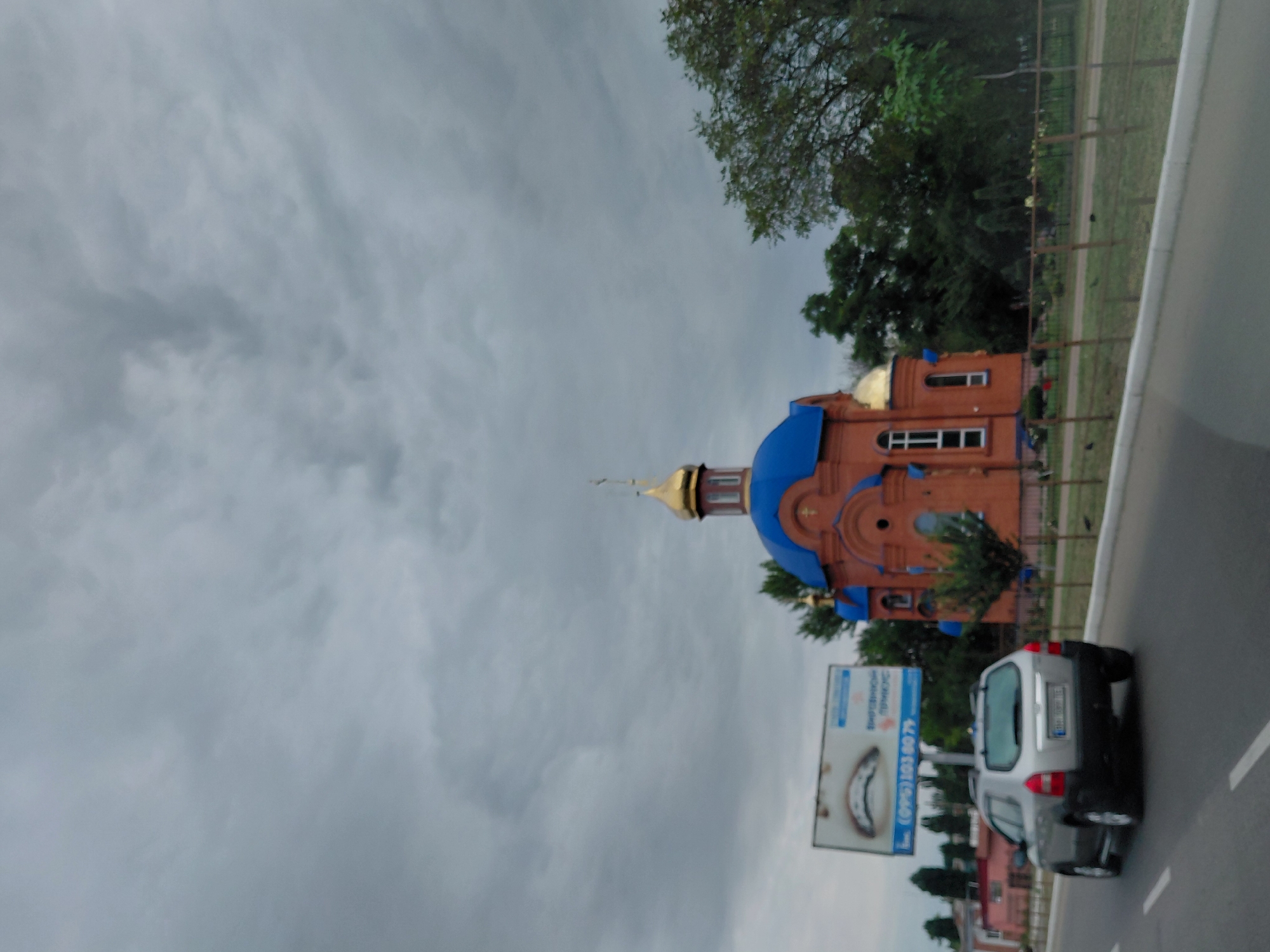 An overcast day scene captures a silver SUV driving away from the camera on a paved road. To the left of the road is a billboard with an image of teeth and text. Further left, a building with pink walls is visible. To the right of the road is a brick church with a blue roof and golden dome, surrounded by trees. The sky is filled with dark, heavy clouds.