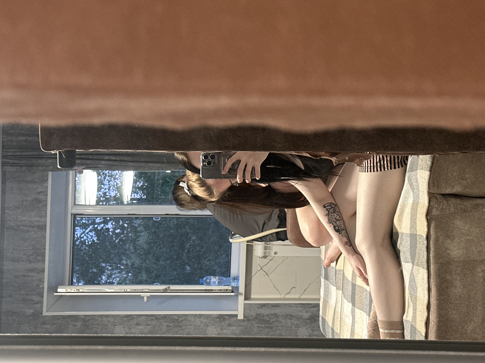 A person is taking a mirror selfie in a bedroom. The person is sitting on a bed covered with a checkered blanket and a brown valance. They are wearing light-colored socks with white stripes and dark-striped shorts. A tattoo is visible on their left arm. In the reflection, a second person is partially visible, sitting on a chair near a window. The window shows trees and what appears to be the sky. A blue water bottle is on the windowsill. The walls have a textured gray finish. A large, blurry, tan object, possibly a piece of furniture or a curtain, is in the foreground, obscuring a portion of the reflection. The lighting suggests it is daytime or early evening.