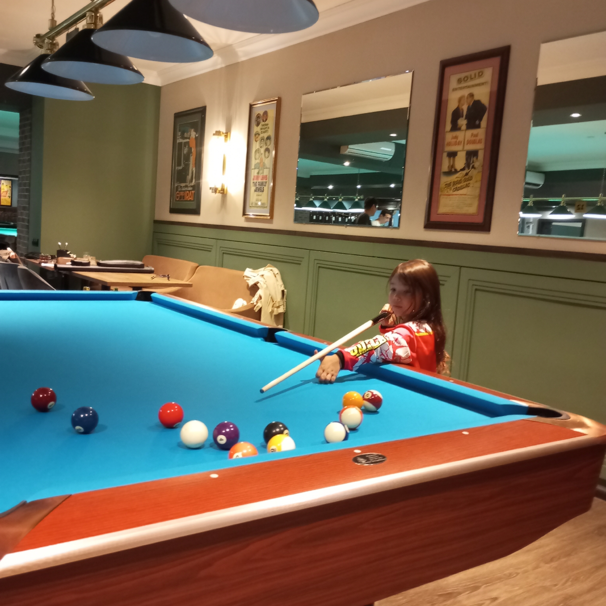 The image depicts an indoor billiards room. A young girl, wearing a red long-sleeved shirt with white graphics, is positioned at a blue-felt pool table, holding a cue stick. She is looking towards the viewer, with her body partially obscuring the pool table. Several billiard balls of various colors (red, blue, white, purple, orange, and yellow) are scattered across the table. In the background, there are framed pictures on the wall, a mirror reflecting ceiling lights and a few individuals, and cushioned seating. The overall environment suggests a recreational facility. There is no indication of the city of Izhevsk, Russia, within the image itself.