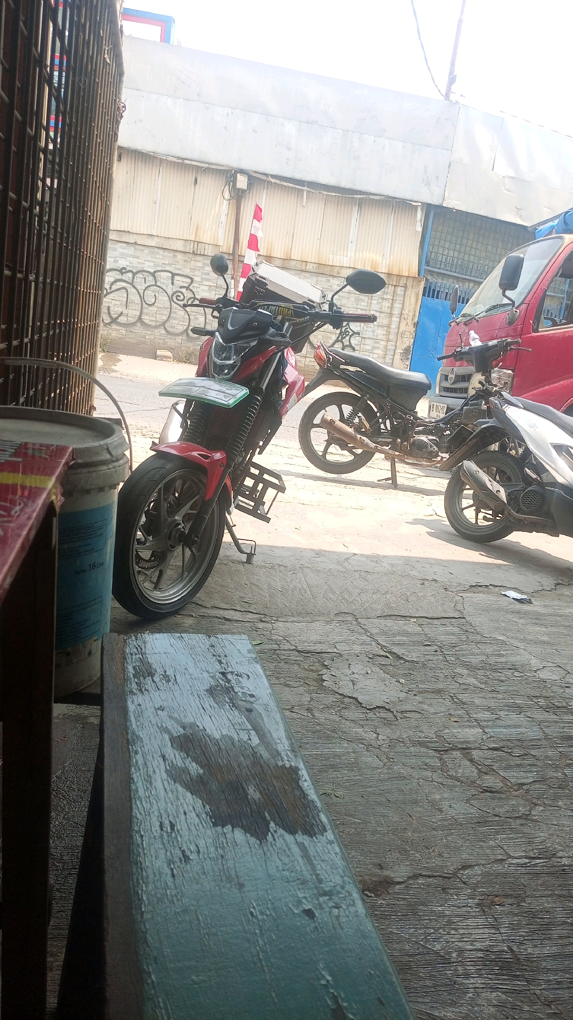 The image depicts an outdoor scene in what appears to be a commercial or residential area, possibly a street or alleyway, in Kadu, Indonesia. In the foreground, a weathered, painted wooden surface, possibly a bench or table, shows signs of wear and moisture. To the left, a large, light-colored plastic bucket with blue lettering is visible, partially obscured.
Dominating the mid-ground are several parked motorcycles. A red and black sport motorcycle is prominently positioned, its front wheel turned slightly to the left. Behind it, a black motorcycle is visible with its kickstand down. Further to the right, a lighter-colored scooter or motorcycle is parked.
In the background, a corrugated metal building is situated, with a portion of a red vehicle, likely a truck or van, visible to the right. A pole with red and white striped markings stands near the building, possibly indicating a safety zone or marker. Graffiti is present on a lower section of the building. The ground surface is paved with concrete or similar material, showing cracks and variations in texture. The overall lighting suggests it is daytime, with sunlight casting shadows. There are no people clearly visible in the frame.