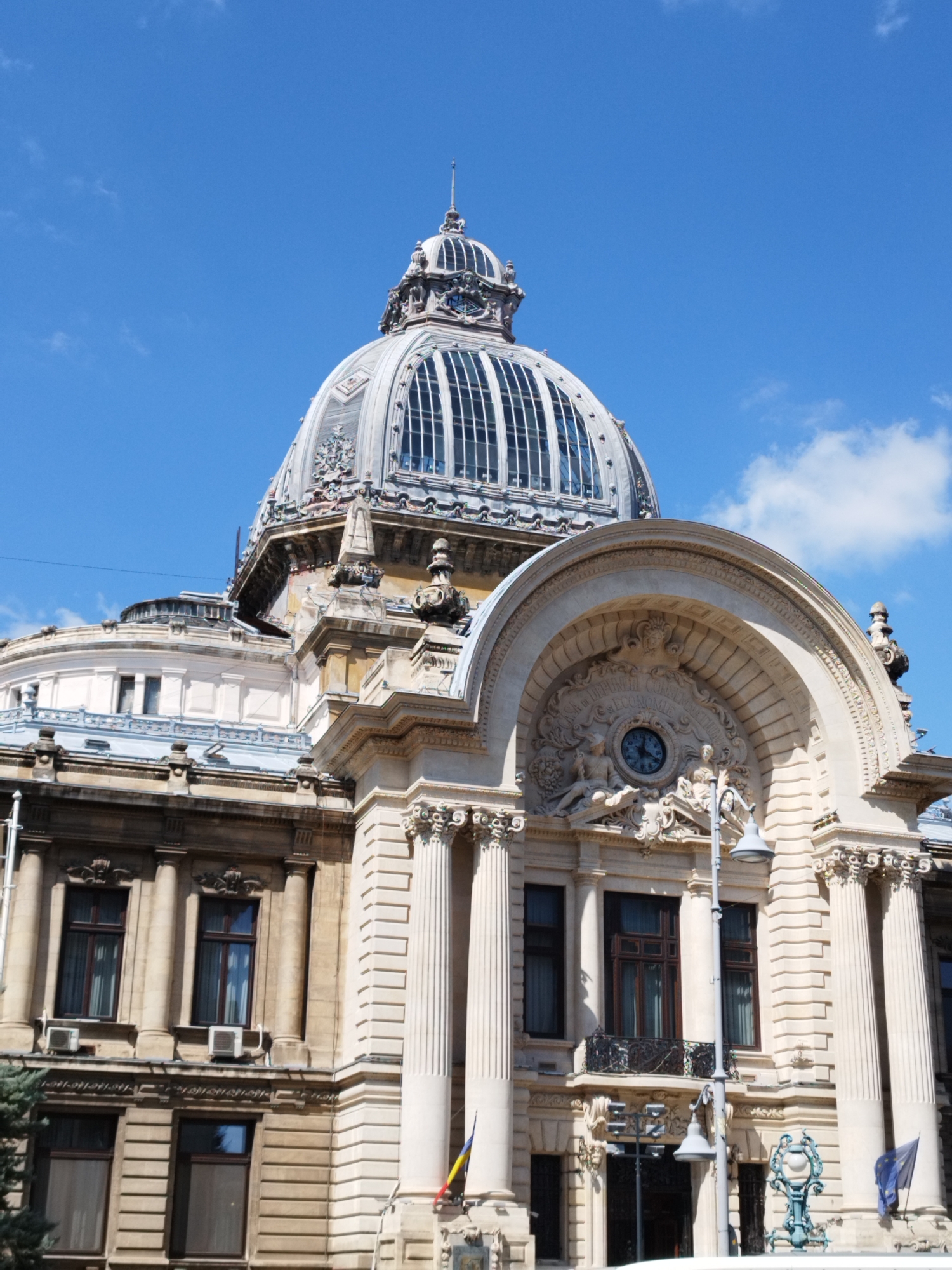 The image shows the Palace of the Deposit and Consignments Fund, located in Bucharest, Romania. The building features a prominent dome and a neoclassical facade with columns and ornate architectural details. A clock is visible on the facade. The sky is blue with some clouds. The image is taken from a low angle, looking up at the building.