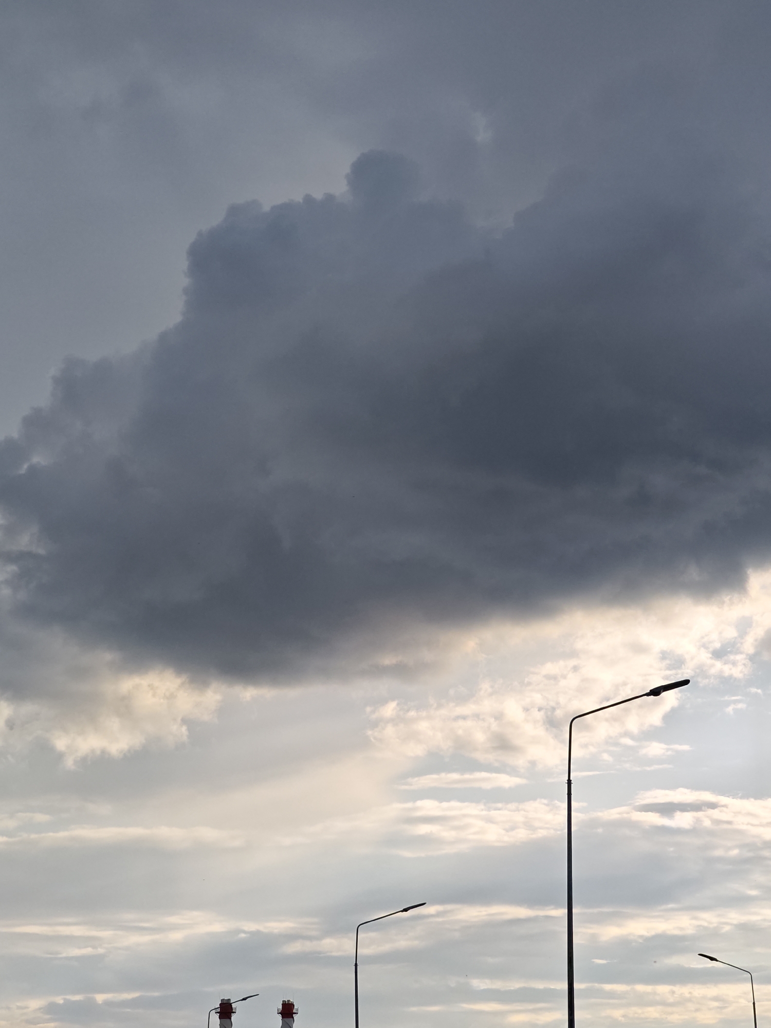 The image depicts a cloudy sky with a large, dark cloud formation dominating the upper portion. Below the dark clouds, lighter, wispy clouds are visible, illuminated by diffused sunlight. Silhouetted against the sky are several streetlights. A prominent streetlight stands in the middle right of the frame, with a thinner streetlight to its left and another to the far right. In the lower portion of the image, two industrial chimneys with red and white banding are visible, also silhouetted. The scene appears to be an outdoor setting in the city of Shcherbinka, Russia, likely during daylight hours given the sky's appearance, though the heavy cloud cover suggests overcast or approaching stormy weather. There are no people or animals visible in the frame.