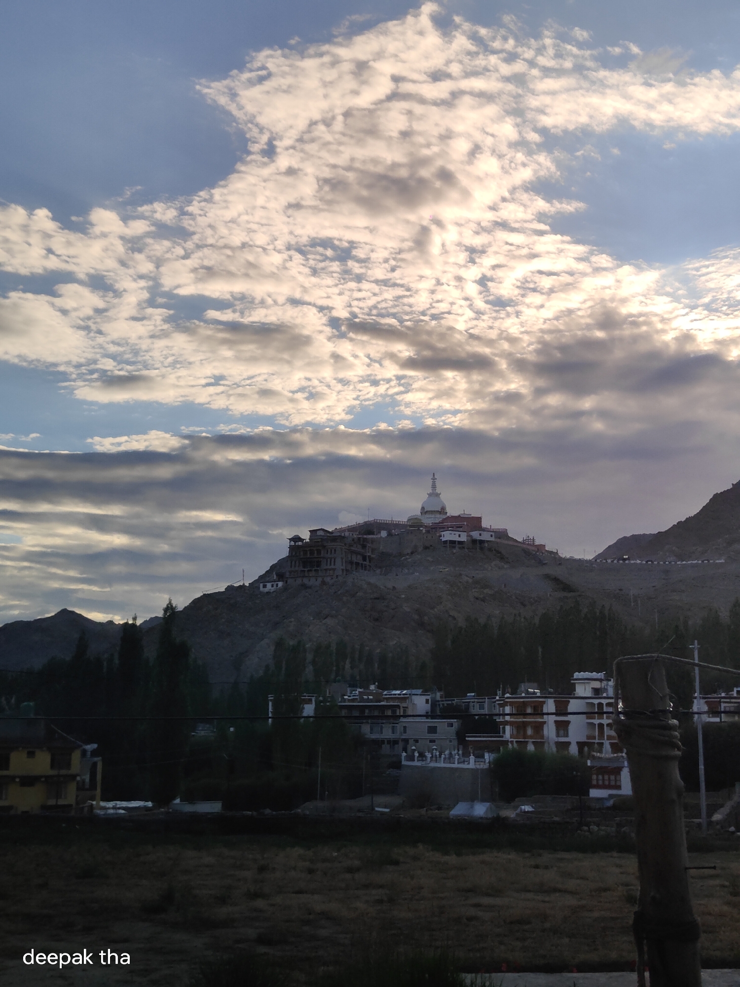 The image captures a wide shot of the Leh Palace and Shanti Stupa complex situated on a hill overlooking the town of Leh, India. The sky is dramatic, filled with a mix of bright, white, and dark gray clouds, suggesting either sunrise or sunset. The structures on the hill appear to be a mix of old stone buildings and newer white and red-colored constructions, including a prominent white stupa with a golden spire. In the foreground, the lower part of the town is visible, with several buildings featuring distinct white and red architectural elements. Trees line the base of the hill. The foreground also shows a dry, grassy field and a wooden post with a rope. The overall impression is one of a serene, mountainous landscape with significant cultural and architectural landmarks.