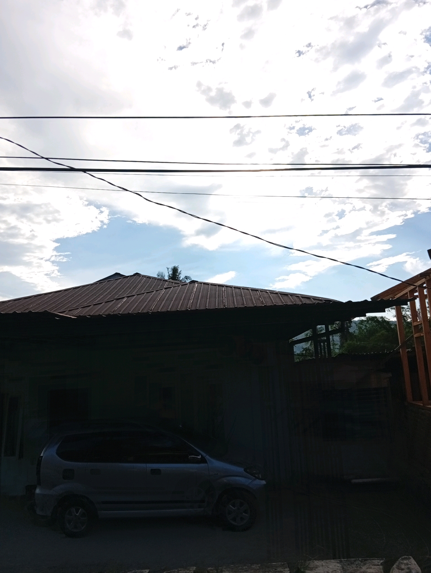 An over-the-shoulder, eye-level shot captured outdoors in the city of Gunungsitoli, Indonesia, shows a silver minivan parked on a paved surface in front of a house. The house features a dark, corrugated metal roof and its facade is mostly in shadow. Overhead, power lines traverse the frame against a bright, partly cloudy sky. To the right of the house, a portion of a structure with orange wooden beams is visible. The background reveals a dense, green forest and a hillside under a hazy sky.
