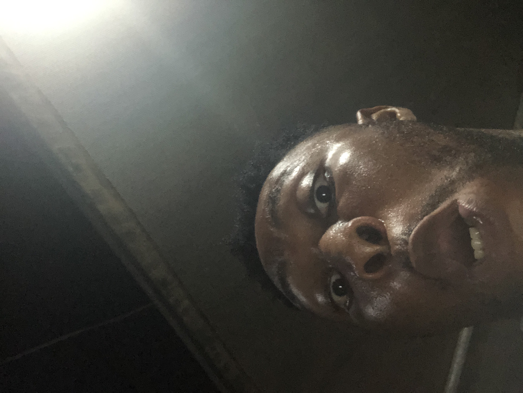 A close-up, low-angle shot depicts a person with dark skin. The person's face is angled upwards and towards the camera. Sweat or moisture is visible on their forehead and face. The lighting in the scene is uneven, with a bright light source in the upper left corner that casts a strong glow. The background is dark and mostly obscured, with some indeterminate dark objects or structures visible in the lower left. The person's eyes are wide open, and their mouth is slightly open, revealing their teeth. The scene appears to be indoors in an unknown location.