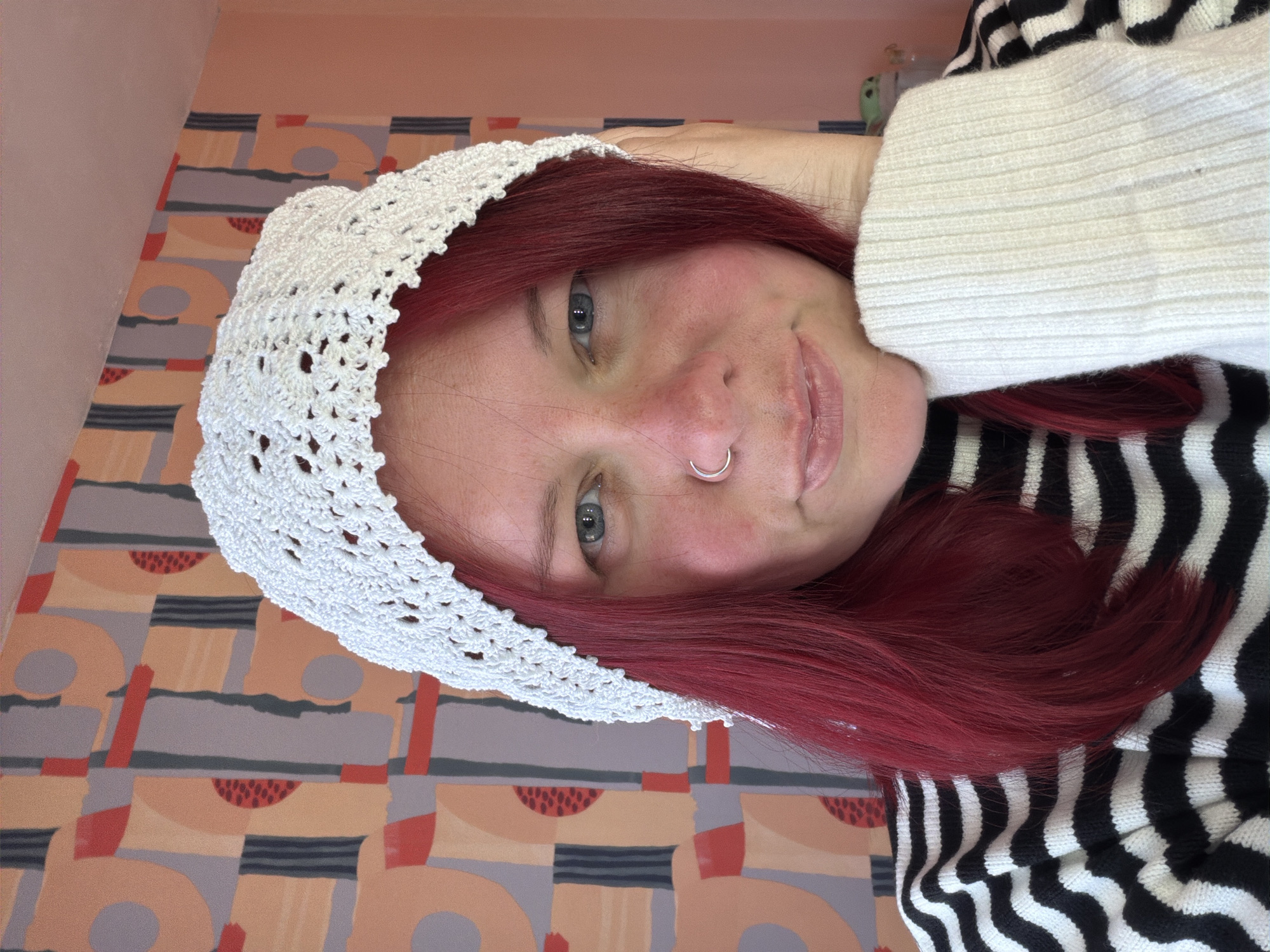 A woman with red hair, blue eyes, and a nose ring is wearing a white crocheted hat. She is also wearing a white sweater and a black and white striped garment underneath. The background is a wall with a patterned design of peach, grey, red, and black shapes. The image appears to be taken indoors. There is no evidence to suggest that the location is the city of Chaikovsky, Russia.