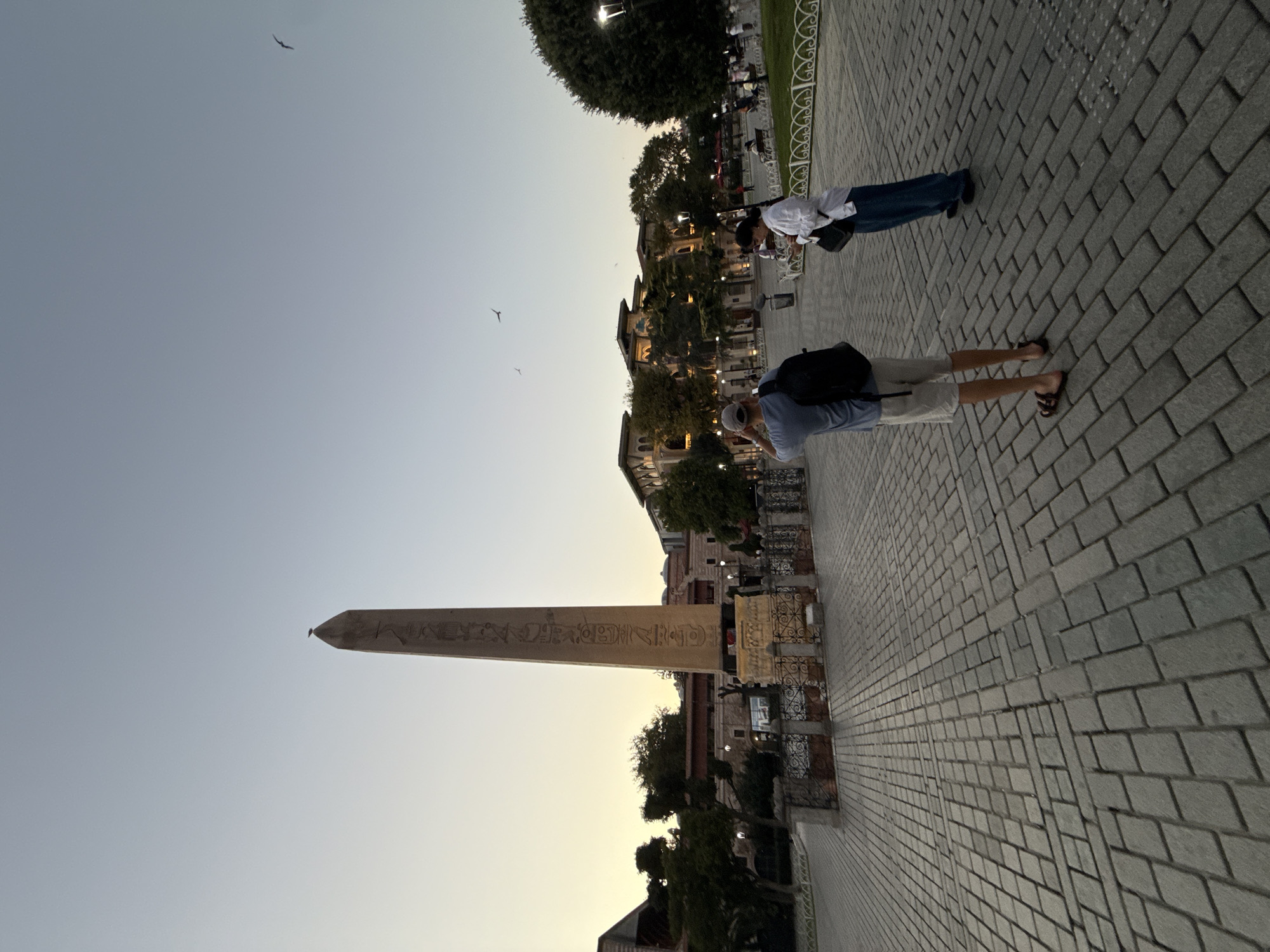The image captures a view of the Sultanahmet Square in Istanbul, Turkey, during the late afternoon or early evening. In the foreground, a paved stone pathway stretches out, leading the eye towards the main subjects. Two individuals are standing on the pathway, facing away from the camera. The person on the left is a male wearing a blue t-shirt, light-colored shorts, and sandals, carrying a black backpack. He is holding a phone or camera up to his face, appearing to take a photograph. To his right stands a female in a white button-down shirt and blue pants, also holding a phone and looking down at it.
Dominating the mid-ground is the ancient Egyptian obelisk, part of the Hippodrome of Constantinople. The obelisk stands tall and slender, with visible hieroglyphics carved into its surface. It is set within a low, decorative stone enclosure. Behind the obelisk and to the right, several buildings with ornate architecture are illuminated, suggesting the presence of historical sites and establishments within the square. Lush green trees dot the landscape, interspersed with streetlights and park benches. In the sky, a few birds are visible, and the overall lighting indicates a soft, natural light as the day transitions into evening. The atmosphere appears to be calm, with other visitors visible in the background, enjoying the open space.