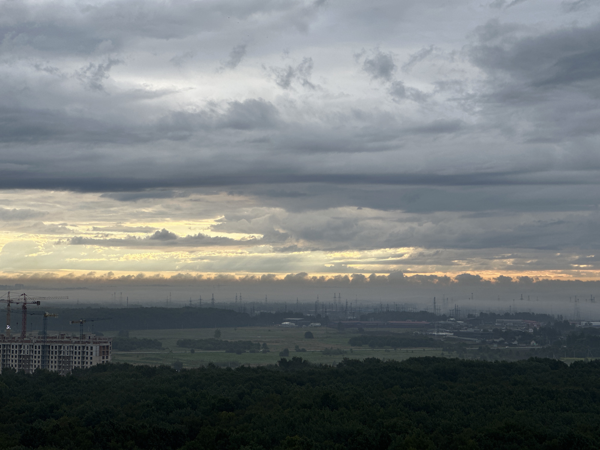 This image captures a wide-angle view of Kudrovo, Russia, under a cloudy sky. In the foreground, a dense forest of dark green trees occupies the bottom portion of the frame. Behind the forest, a large, multi-story building under construction is visible on the left, with several construction cranes surrounding it. Further in the distance, a hazy, industrial landscape stretches across the horizon, characterized by numerous power lines and structures. The sky is filled with layers of grey clouds, with a band of lighter, yellowish clouds near the horizon, suggesting either sunrise or sunset, with some rays of light breaking through. The overall scene depicts a developing urban area adjacent to a natural landscape, with an industrial zone in the background.