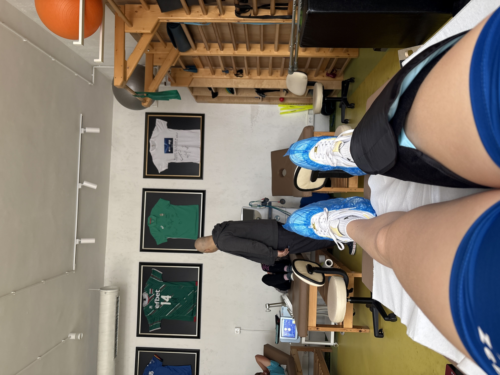 The image, taken from a first-person perspective, captures an indoor scene within what appears to be a physical therapy or rehabilitation clinic, possibly in Sofia, Bulgaria. The viewer's legs are extended forward, resting on a white-sheeted examination table. The person is wearing blue athletic shorts and white sneakers covered with disposable blue shoe covers. A black brace or wrap is secured around the upper thigh and knee area of the right leg, suggesting injury recovery or therapy.
The room features white walls and a light yellow floor. In the midground, a bald man in a dark hoodie and pants stands near various medical and rehabilitation equipment. This includes a beige therapy table, a wheeled stool, and a machine with a digital screen, possibly used for electrotherapy or diagnostics. An extensive wooden shelving unit fills the background, holding various exercise and therapy tools such as resistance bands and small balls. An orange exercise ball is suspended from a ceiling-mounted rack.
The left wall is adorned with several framed sports jerseys, indicating a connection to athletics. Visible jerseys include a white one with the "OMV" logo and signatures, two green ones (one with the "efbet" logo and the number "14," and another with the "asics" logo), and a partially visible blue jersey with a "DB" logo. The room is illuminated by overhead and strip lighting mounted on the walls. A tall, white air purifier stands near the framed jerseys. The overall setting, combined with the brace on the leg, suggests the person is undergoing a rehabilitation session.