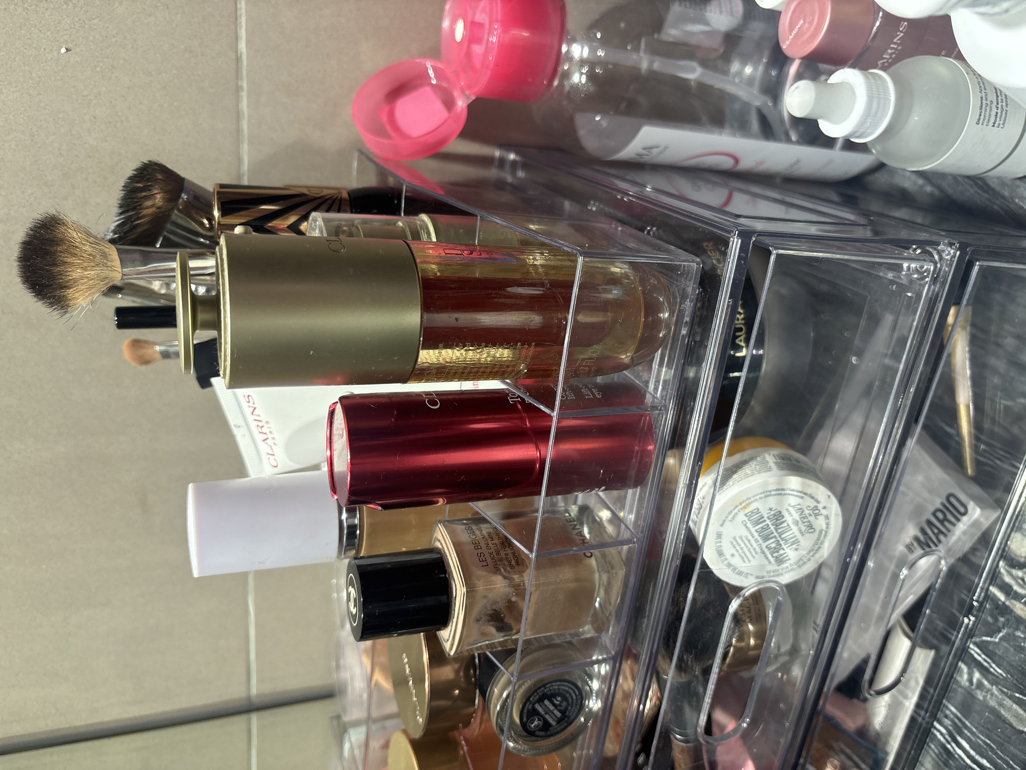 The image displays a close-up, eye-level view of a clear acrylic makeup organizer filled with various beauty products. The organizer is situated on a reflective surface, likely a countertop, with a tiled wall in the background.
Prominent items include several bottles of skincare and makeup, brushes, and other cosmetic containers. A large gold-capped bottle with a clear liquid is centrally positioned. To its left, a metallic red cylindrical container is visible. A clear bottle labeled "CHANEL LES BEIGES" containing a light-colored liquid is to the left of the red container, and a smaller dark-capped glass bottle is to the left of that. In the foreground, a white jar with yellow lettering, which appears to be a body cream, is visible within a drawer of the organizer, along with a tube or package labeled "DR MARIO."
Further to the right, a clear bottle with a pink pump dispenser is prominent, alongside other bottles of varying sizes and colors, some of which appear to be skincare serums or toners. Makeup brushes with visible bristles are standing upright in a holder behind some of the bottles.
The lighting appears to be artificial, casting subtle reflections on the surfaces. The overall impression is one of an organized personal collection of beauty products.
There is no information in the image to suggest a location in Dubai, United Arab Emirates.