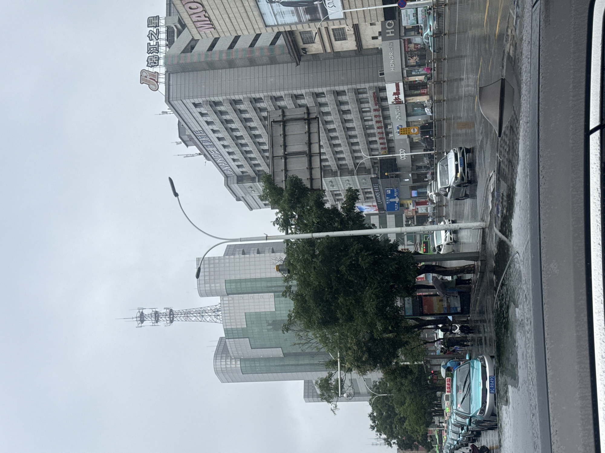 The image depicts a street scene in Qinhuangdao, China, on a rainy day. The sky is overcast and the wet pavement reflects the surrounding buildings and streetlights. Multiple lanes of traffic are visible, with several cars and a line of teal-colored taxis in the foreground. People are seen holding umbrellas and walking along the sidewalks, some under covered awnings of shops. Buildings of varying architectural styles line the street, including modern high-rises with glass facades and older structures with visible signage. A prominent cell tower stands tall against the sky, and a large billboard is affixed to a building. The overall impression is of an active urban environment experiencing inclement weather.