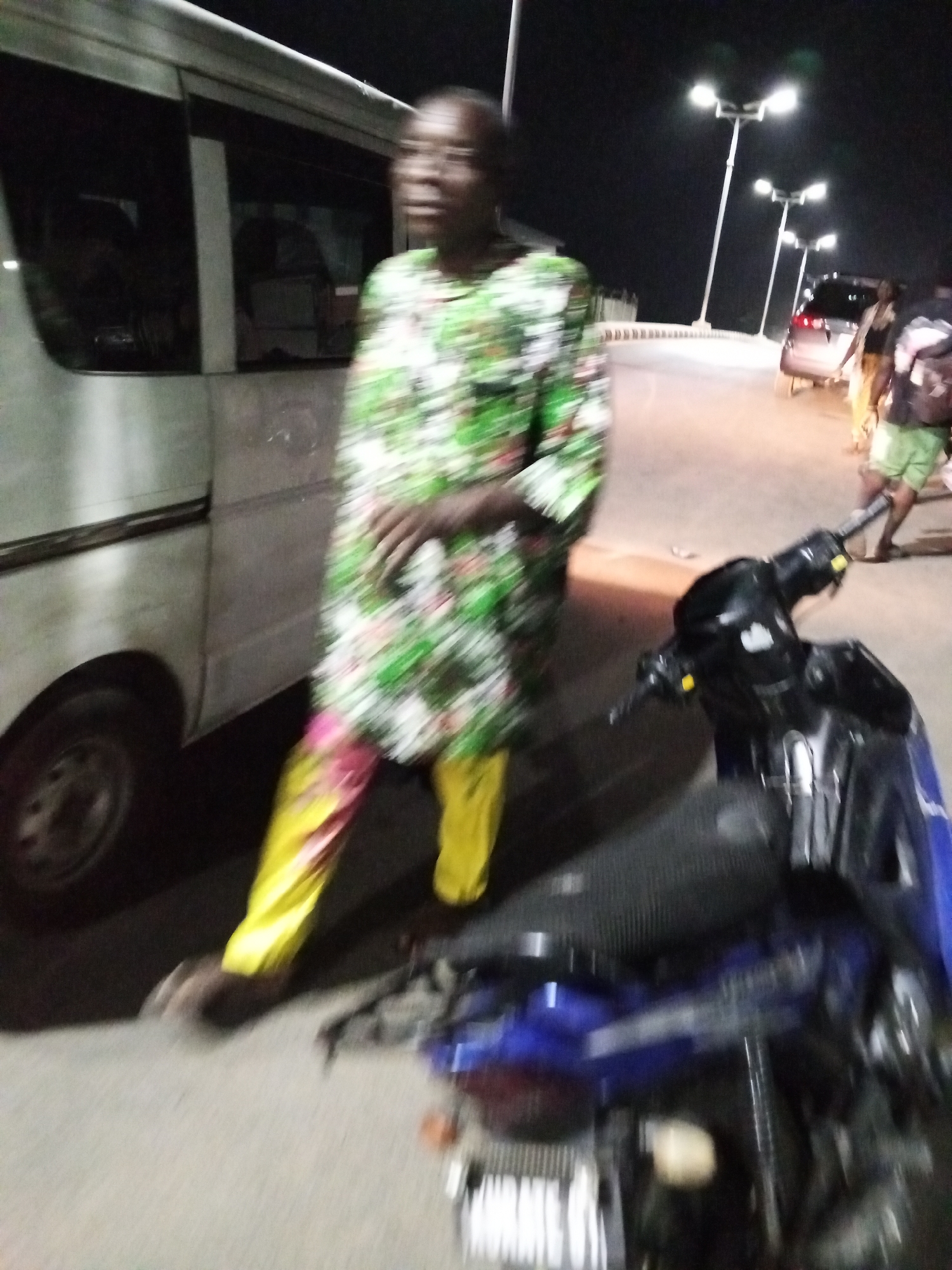 In Adankolo, Nigeria, a blurred image captures a person in a green and white floral patterned top and yellow pants standing beside a white van. The person is facing forward with their arms crossed. To the right, a parked motorcycle is visible, its details somewhat indistinct. In the background, street lights illuminate a road with at least one car and several other individuals on the side of the road.