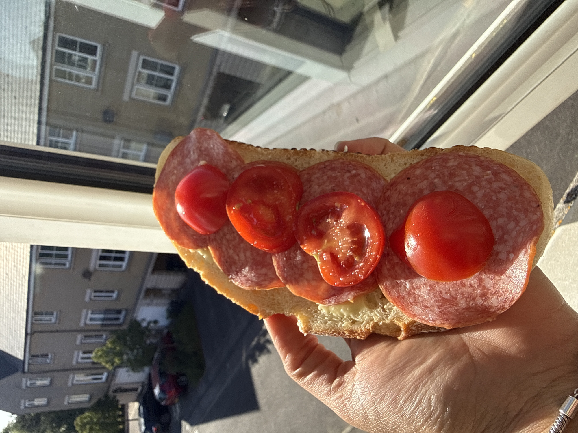 A sunlit photograph depicts a hand holding a sandwich. The sandwich consists of a bread roll topped with slices of salami and halved cherry tomatoes. The background shows an outdoor scene, likely viewed through a window. There is a building facade with multiple windows visible, and a street scene is partially visible below, with parked cars and greenery. The photograph was taken in Great Yarmouth, United Kingdom.