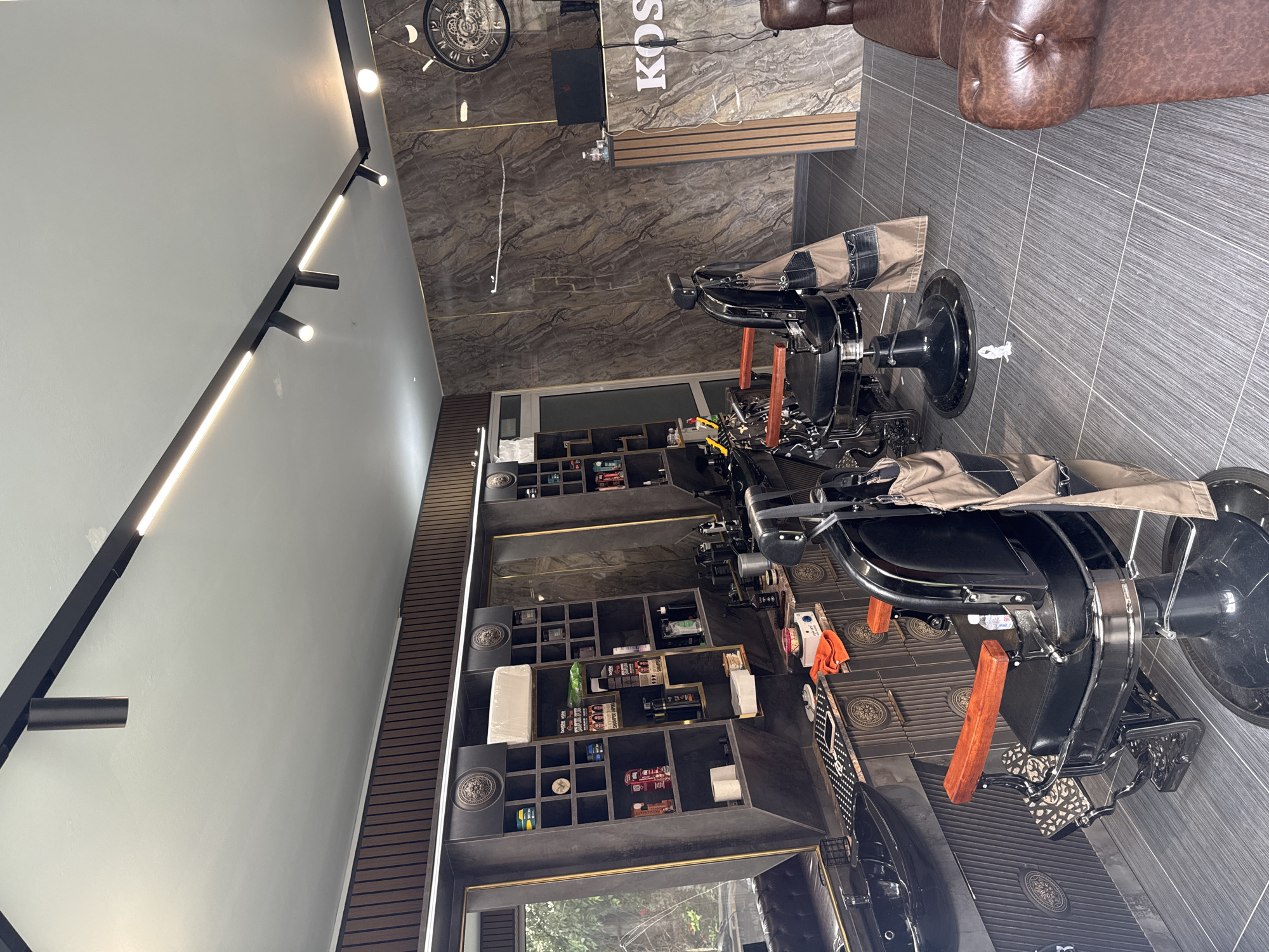 This media file captures the interior of a modern and well-appointed barbershop or salon, likely located in Varna, Bulgaria. The space is empty of people, suggesting either opening or closing hours, or a lull in business.
The shop features a sleek design with contrasting materials. The ceiling is light grey with black track lighting and integrated LED strips, running along a dark structural element. The floor is covered in dark grey tiles, some with subtle patterns, particularly around the bases of the barber chairs.
Two prominent black barber chairs with tan fabric covers or capes are visible, both facing away from the camera. These chairs have classic designs with wooden armrests, positioned on the patterned floor tiles. Further back, a comfortable-looking brown tufted leather sofa provides seating for clients awaiting service.
Along one wall, a striking shelving unit is built into dark wooden slats, framed with gold accents. This unit displays various hair care products, towels, and other salon supplies, neatly arranged within individual compartments. At the back of the shop, a dark marble-patterned counter serves as a reception or workspace. On this counter, a laptop and several bottles are visible, and the partial text "KOS" can be seen, likely part of the business's name. A large, dark, ornate wall clock with Roman numerals hangs above the reception area.
Natural light filtering through a window or glass door at the far end indicates it is daytime. The overall impression is one of cleanliness, professional styling, and an inviting atmosphere.