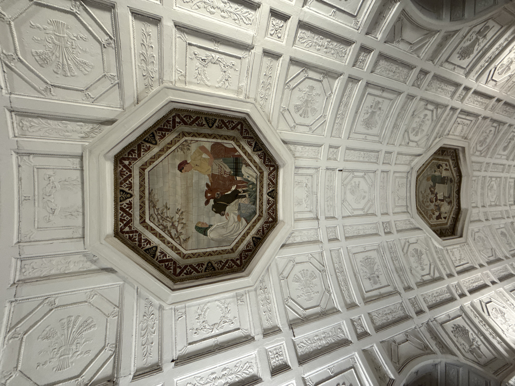 The image shows an ornate, coffered ceiling in a building, likely in Moscow, Russia. The ceiling is primarily white and features intricate plasterwork in relief. The geometric patterns of the coffers are varied, including octagonal and square panels, many of which are decorated with floral motifs, stylized wheat stalks, and urns.
Dominating the center of the image is a large octagonal panel containing a painted fresco. The fresco depicts a scene with two figures and a cow. One figure, dressed in a yellow garment and a red cap, appears to be interacting with the cow. Another figure, dressed in white and a green head covering, is also present. The fresco is bordered by a decorative frame with a geometric pattern of red, brown, and black.
To the right of the main fresco, a smaller octagonal panel is visible, also containing a fresco. This smaller fresco appears to depict a similar scene with human figures and possibly an animal or object, though it is less detailed in the image.
The overall impression is one of grandeur and historical significance, suggesting the ceiling is part of a public building, a museum, or a historical residence. The lighting appears to be indoor lighting, and no specific time of day or weather conditions are discernible. There is no visible text in the image.