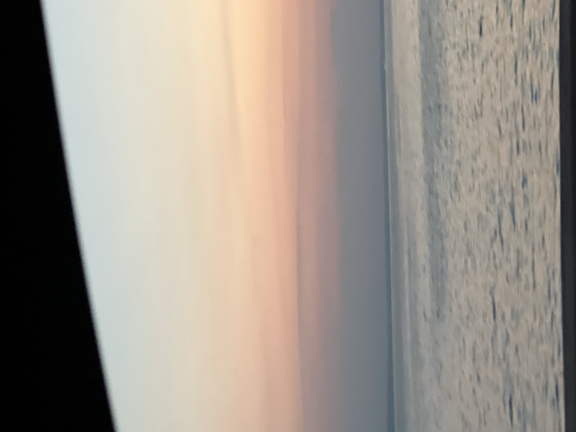 A body of water, possibly a river or lake, is shown under a sky with a sunset. The sky transitions from a pale yellow at the top to pink and then a soft purple-gray near the horizon. A dark, out-of-focus band is visible at the very top of the image, obscuring a portion of the sky. The water's surface is calm, with gentle ripples reflecting the sky's colors. The scene appears to be devoid of any people or man-made structures, giving a sense of tranquility. The location in Chaikovsky, Russia, is not explicitly indicated by any visual cues in the image itself.