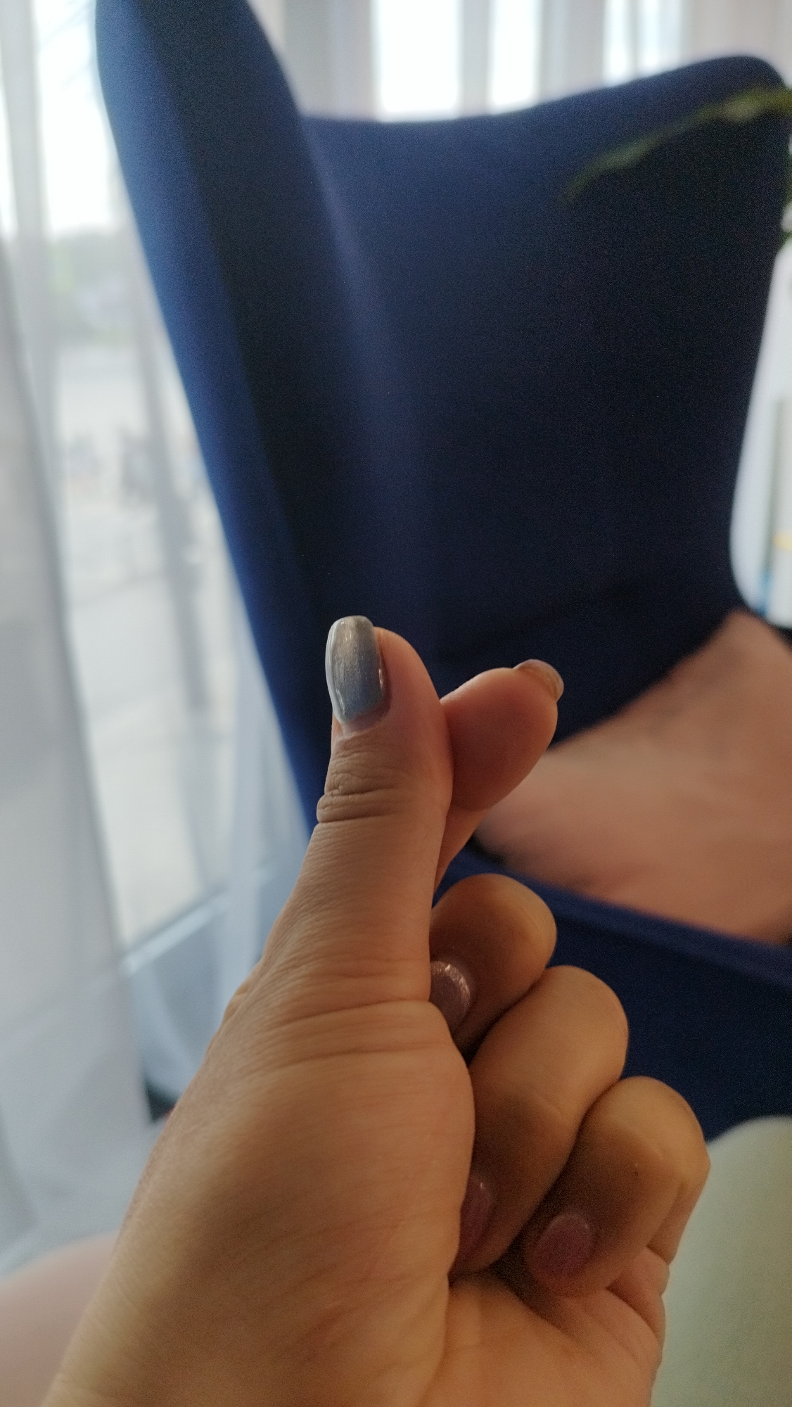 A close-up, low-angle, medium shot captures a person's left hand making a finger heart gesture in front of a blurred blue chair and a window. The person's thumb and index finger are touching, forming the shape of a heart. The fingernails are painted with a shimmery light gray polish on the thumb and index finger, and a light pink polish on the other visible fingernails. The background is softly out of focus, with the dark blue fabric of the chair dominating the right side of the frame and a bright window with sheer curtains visible on the left. The lighting is natural and diffused.