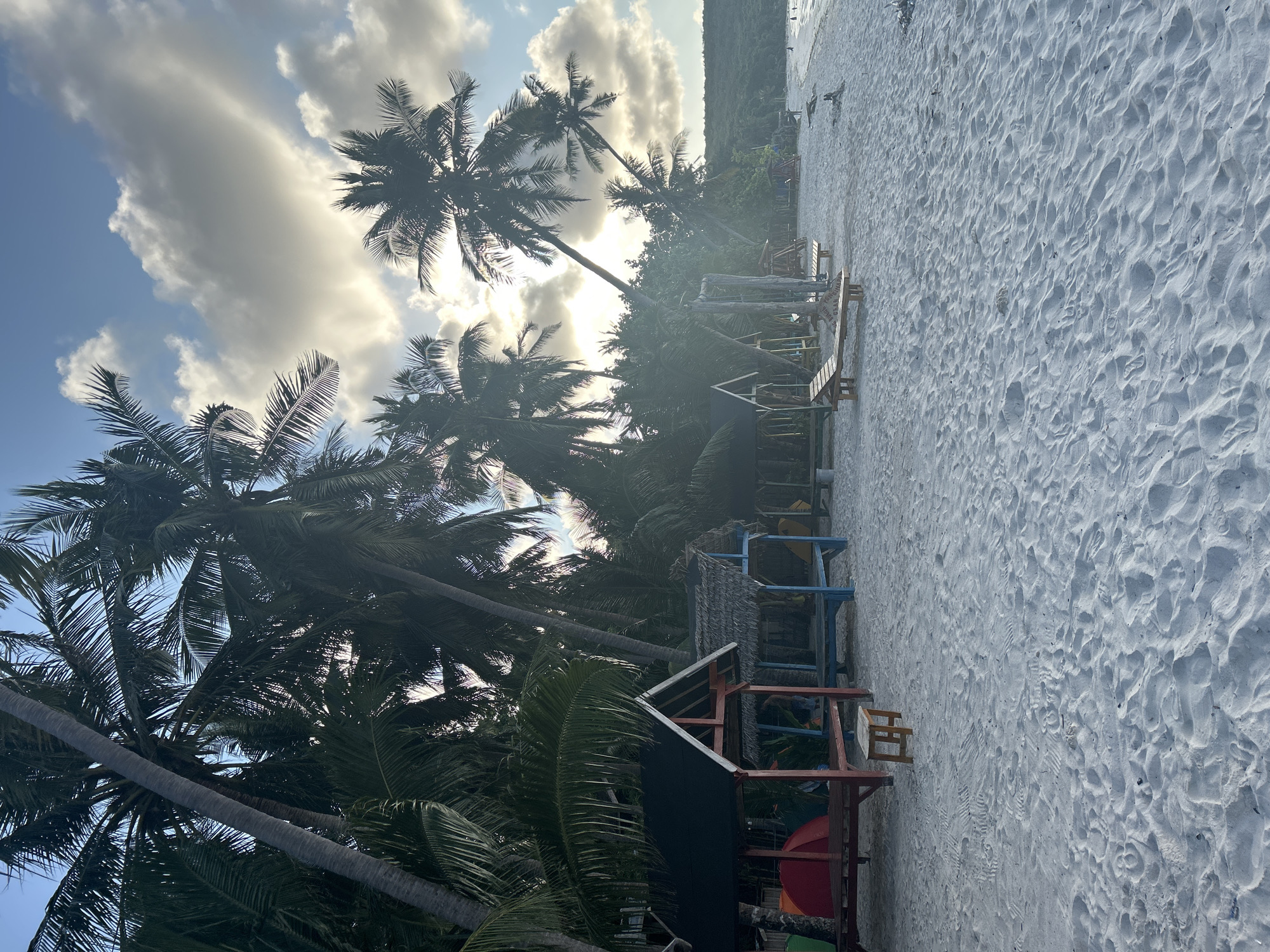 The image depicts a tropical beach scene with white sand, palm trees, and several small huts. The huts are made of wood and thatch, with one having a red and black roof and another a blue base and thatched roof. There are also lounge chairs and swings on the beach. In the background, lush green vegetation lines the shore, leading up to a hillside. The sky is partly cloudy with the sun shining through, creating a bright and atmospheric effect. The scene appears to be a resort or a recreational area. The location is a beach in A R A, Indonesia.