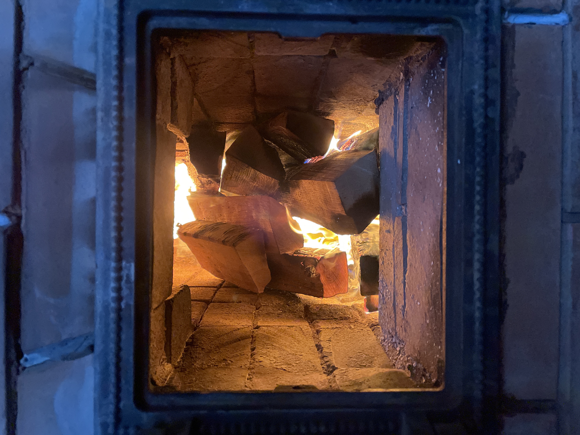 A rectangular wood-burning stove is the primary focus, filled with stacked logs that are actively burning. The interior of the stove is made of firebricks. The environment is dim, with a slight blue hue surrounding the outer edges of the stove's frame. The location is presumed to be Sestroretsk, Russia.