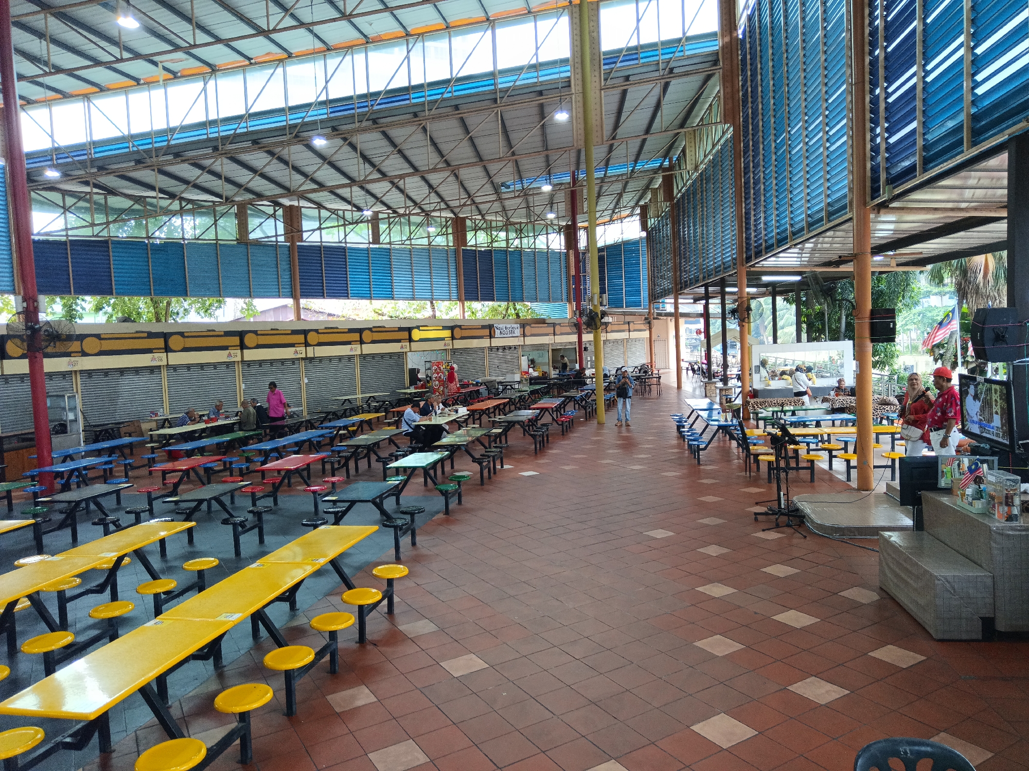 This image depicts an outdoor food court in Kuala Lumpur, Malaysia, under a covered roof supported by metal beams. The roof is partially transparent, allowing natural light to filter through. The food court features numerous long, narrow tables with attached benches, most of which are yellow. The floor is tiled with red and light brown squares. In the background, there are rows of food stalls with rolling metal shutters, some of which are open, revealing glimpses of activity within. Several people are visible in the food court, including individuals seated at tables, walking between them, and standing near the stalls. There are also two individuals in the foreground on the right side of the image, one wearing a red shirt and the other in a red hat and white top, interacting with each other near a television screen and a sound system. Malaysian flags are displayed prominently near the right side of the frame. The overall scene suggests a busy and functional public space for dining.
