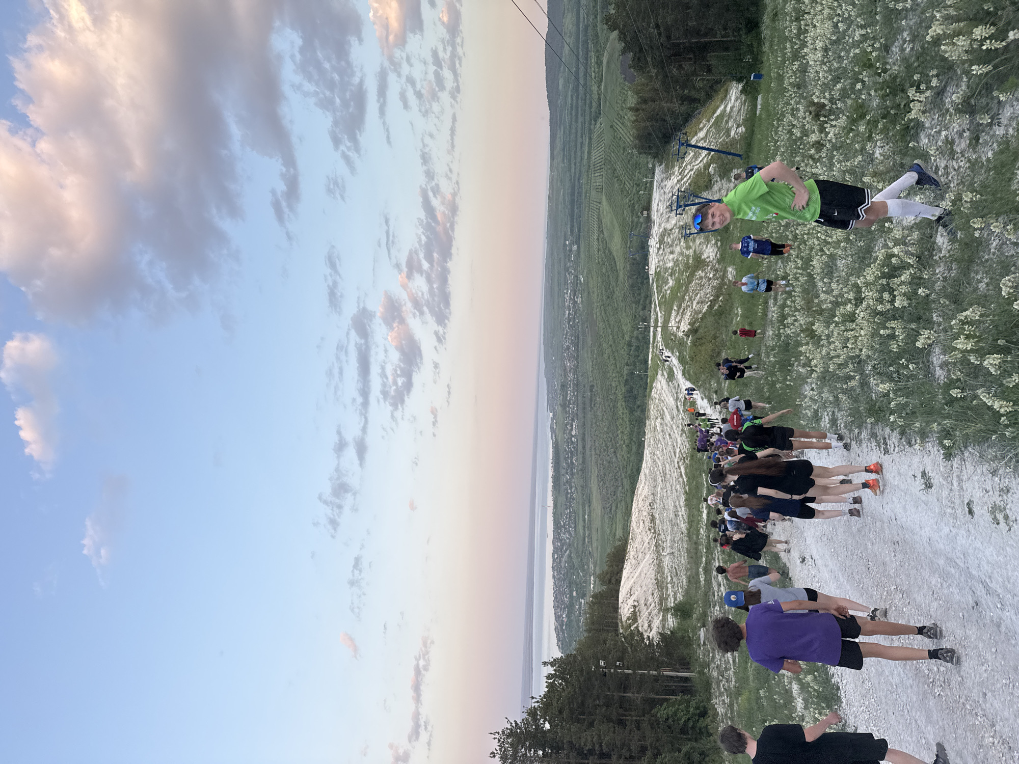 A broad landscape oriented photograph shows a group of people participating in an activity on a hillside, under a colorful sky.
In the foreground, a group of people are lined up on what appears to be a dirt path or clearing. They are dressed in various athletic attire and are seemingly engaged in some form of exercise or activity. The hillside is dotted with patches of white flowers.
In the background, the landscape extends into a valley, possibly overlooking a body of water. The sky is colored with shades of blue, pink, and white, indicating it could be sunrise or sunset. There are visible structures on the hillside, which appear to be ski lifts or cable cars.
The location can be identified as a ski resort on the Chalk Mountains near Khvalynsk, Russia, known for its chalky soil and the Volga River view. The image features participants in the "Chalk Race" ("Меловой забег") event, which is a popular annual sporting event on Chalk Mountain near Khvalynsk.