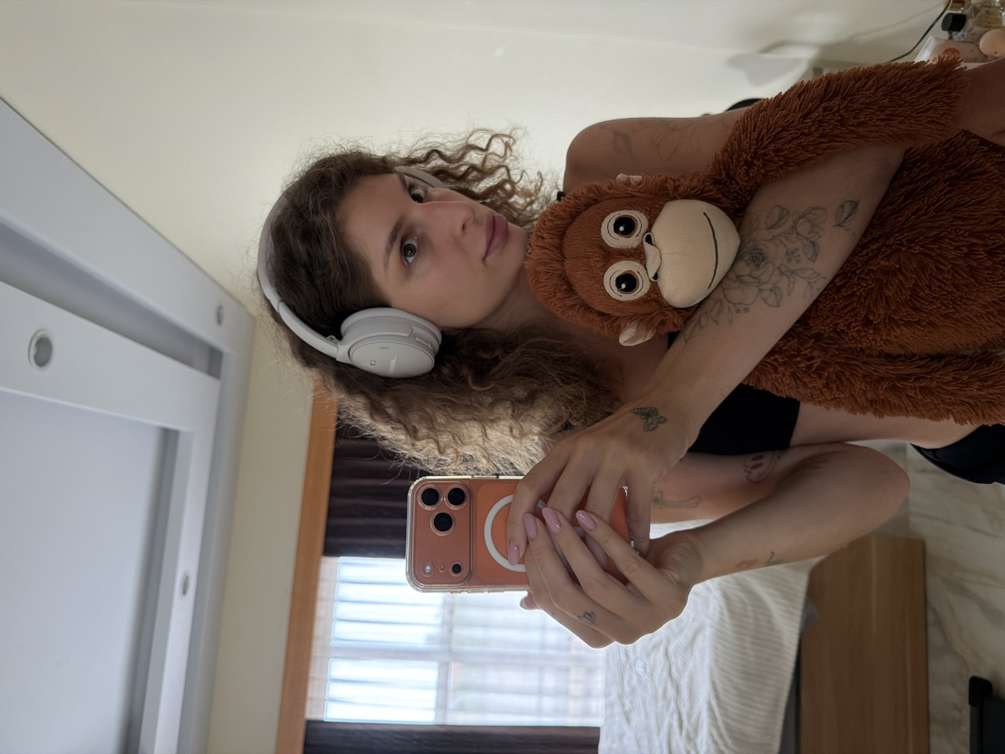 A young woman with curly brown hair and headphones around her ears is holding a mobile phone in her right hand and a plush orangutan toy in her left arm. She is looking at the phone, which is angled towards a mirror, indicating she is taking a selfie. The background shows a bedroom with a bed, curtains, and a ceiling light. The woman has several tattoos visible on her arms. The scene appears to be indoors, possibly in a residential setting in North Kuta, Indonesia.
