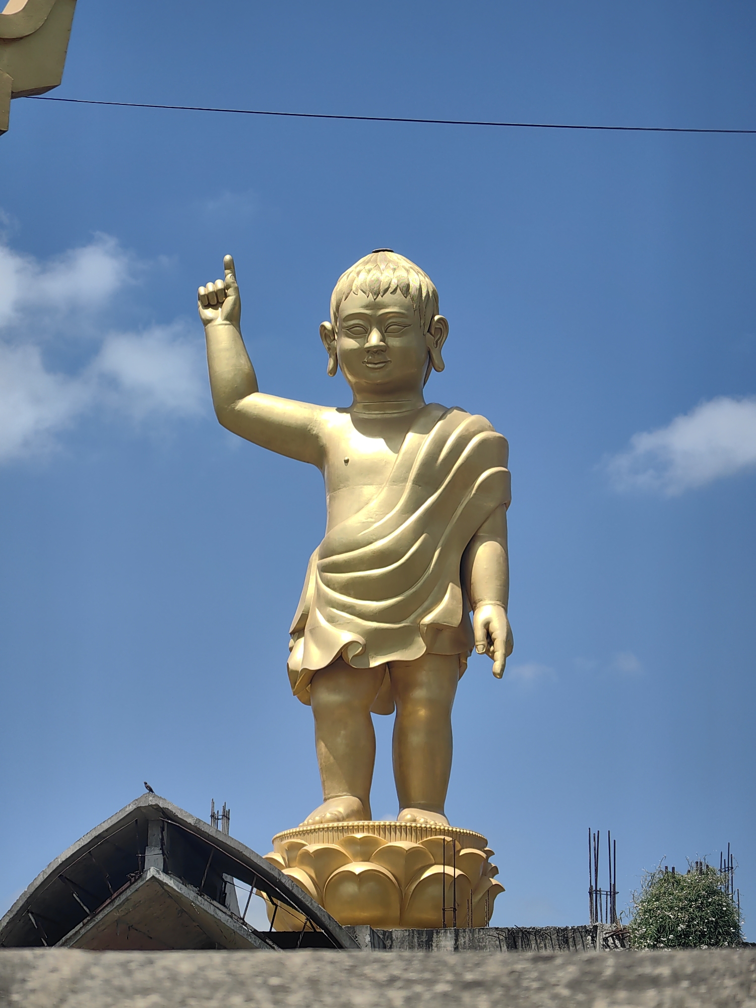 A full-body, low-angle shot captures a golden statue of a young Buddha standing atop a golden lotus pedestal against a bright blue sky with sparse white clouds. The statue, appearing to be made of polished metal, depicts a child with an serene expression, draped in flowing robes. The figure's right arm is raised with the index finger pointing upwards. The base of the statue is supported by a multi-layered golden lotus. In the foreground, a partially constructed building with visible concrete and rebar elements occupies the lower left portion of the image, with a small bird perched on its apex. To the right, more construction materials and a bush with white flowers are visible. The scene is likely located outdoors in a place of religious significance, possibly Lumbini Bazaar, Nepal, given the subject matter and construction elements.