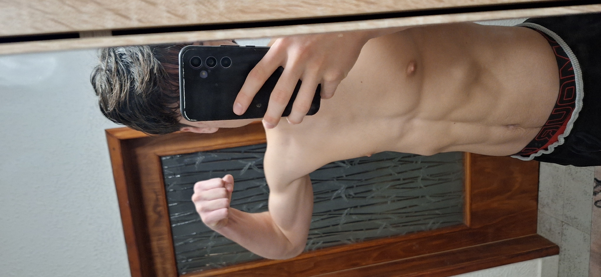 A shirtless person with short dark hair and a muscular physique is taking a selfie in a mirror. They are holding a black smartphone with multiple camera lenses. They are wearing black shorts with a red waistband. The reflection in the mirror shows a window with a dark, textured glass pane and a wooden frame. The background wall appears to be white. The location cannot be determined as Burgas, Bulgaria from the image alone.