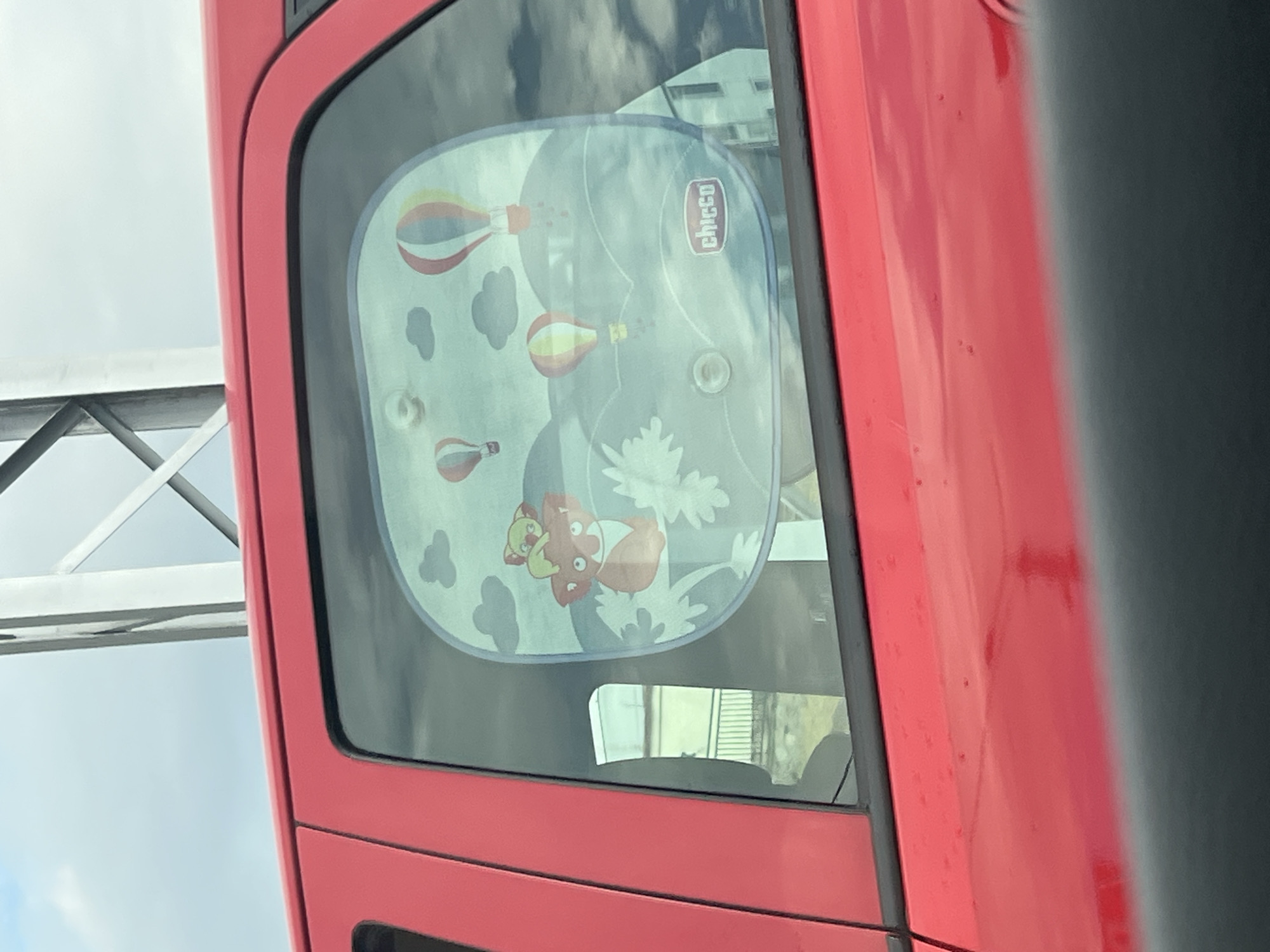 A red vehicle is parked on a bridge with a guardrail visible on the left side. The vehicle's rear side window has a sunshade attached, featuring a cartoon design with hot air balloons, clouds, leaves, and an animal character, along with the "Chicco" logo. The sunshade is partially obscuring the view through the window. The environment appears to be urban, possibly an overpass in Plovdiv, Bulgaria, based on the license plate information. The weather seems overcast. There is no evidence of any people in the frame.