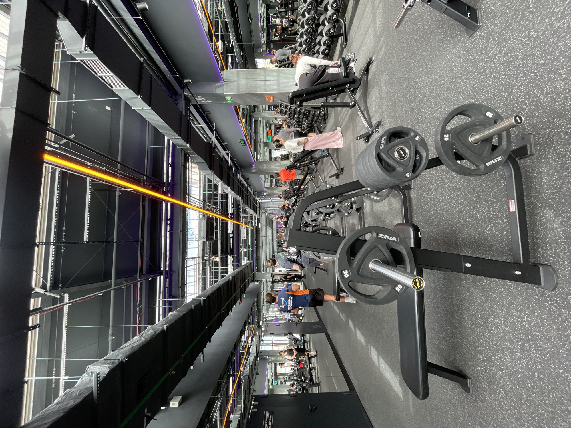 The image shows the interior of a gym, likely in Kazan, Russia, given the "Tatsportobespechenie" branding on the equipment. The space is dominated by various weightlifting machines and free weights. People are working out, with several individuals visible throughout the gym. The environment features a modern, industrial aesthetic with high ceilings, exposed piping, and black-and-gray color schemes. Artificial lighting, including orange linear fixtures, illuminates the space. Weight plates and barbells are prominent in the foreground and background, indicating the gym's focus on strength training.