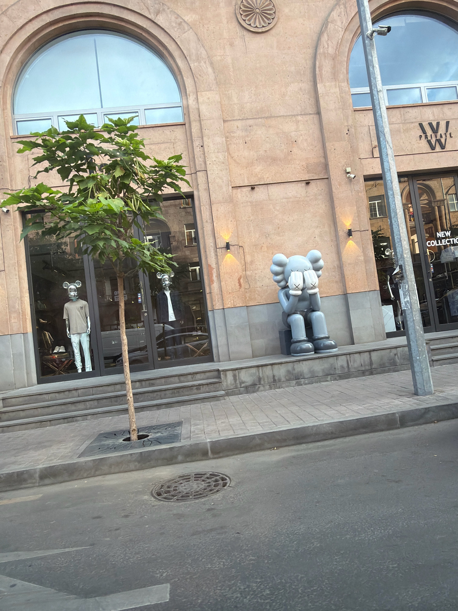 The image captures a street-level view of the facade of a building in Yerevan, Armenia. The architecture is characterized by large arched windows with a light beige stone exterior. A slender tree stands in front of the building, positioned to the left of center.
To the left, two mannequins are displayed in a storefront window. They are outfitted with casual clothing and feature distinctive mouse-like head coverings. One mannequin wears a light-colored t-shirt and pants, while the other is dressed in a darker outfit with a white t-shirt underneath.
To the right of the tree, a large grey sculpture of a seated figure with its eyes covered by its hands is prominently displayed on the building's steps. This figure is a recognizable piece of art, commonly associated with the artist KAWS.
Above the storefronts, a sign with the letters "W | PRIVATE W" is visible. Further to the right, another sign reads "NEW COLLECTION." The building appears to house retail establishments. A light pole with security cameras is situated on the right side of the frame. The street in the foreground shows a manhole cover and the paved road. The overall impression is one of a modern commercial area within the city.