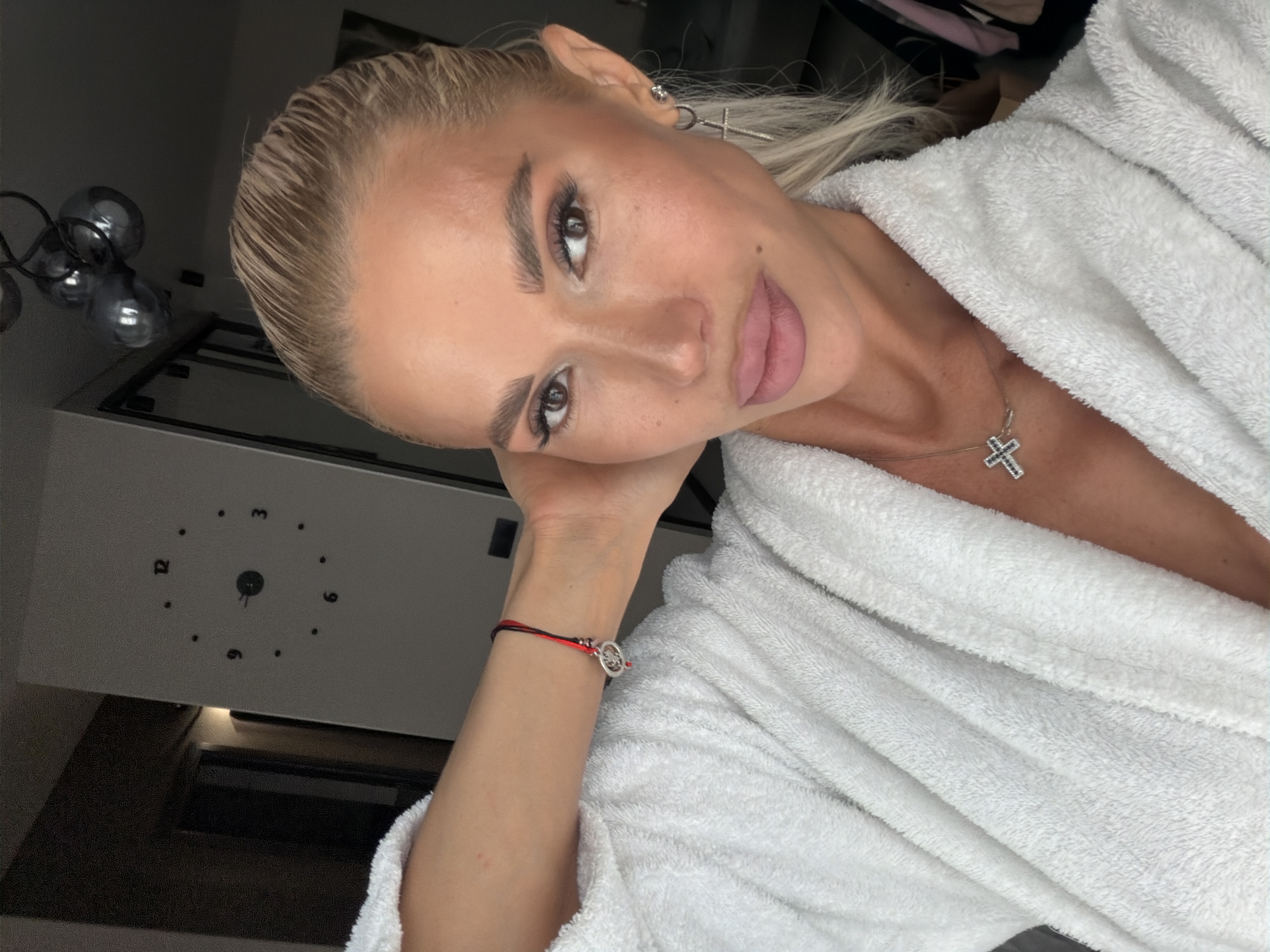 A woman is posing for a selfie. She is wearing a white bathrobe and jewelry, including a necklace with a cross pendant and earrings in the shape of crosses. A bracelet is visible on her wrist. She has light-colored hair and distinct eyebrow makeup. The background includes a lamp with multiple spherical lights and a clock. The location is Unknown, Unknown.