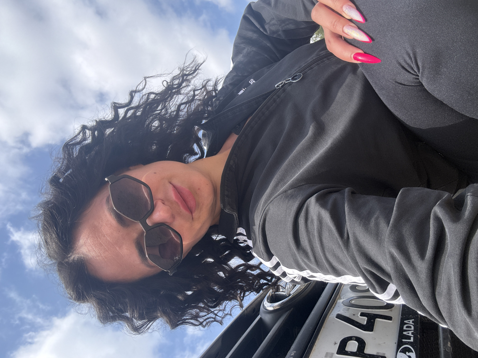 A low-angle, rotated portrait captures a person looking towards the camera against a bright, cloudy sky. The subject, likely female, has dark, curly hair and wears dark, hexagonal-framed sunglasses. Her lips are slightly parted, showing a neutral expression.
She is dressed in a dark, possibly black or charcoal, zippered jacket featuring white stripes on the sleeve, and dark trousers. One hand is visible, resting on her leg, displaying long, pointed nails. Some nails are a vibrant pink/magenta, while others are glittery white with pink tips.
The background is a clear blue sky dotted with scattered white clouds, indicating daytime conditions. A partial view of a dark vehicle is at the bottom of the frame. On its license plate, the characters "P40" and the "LADA" logo are clearly visible, identifying the vehicle as a Lada. The scene is set in Maksimovo, Russia. The image has a casual, outdoor feel, focusing on the subject's appearance and style.