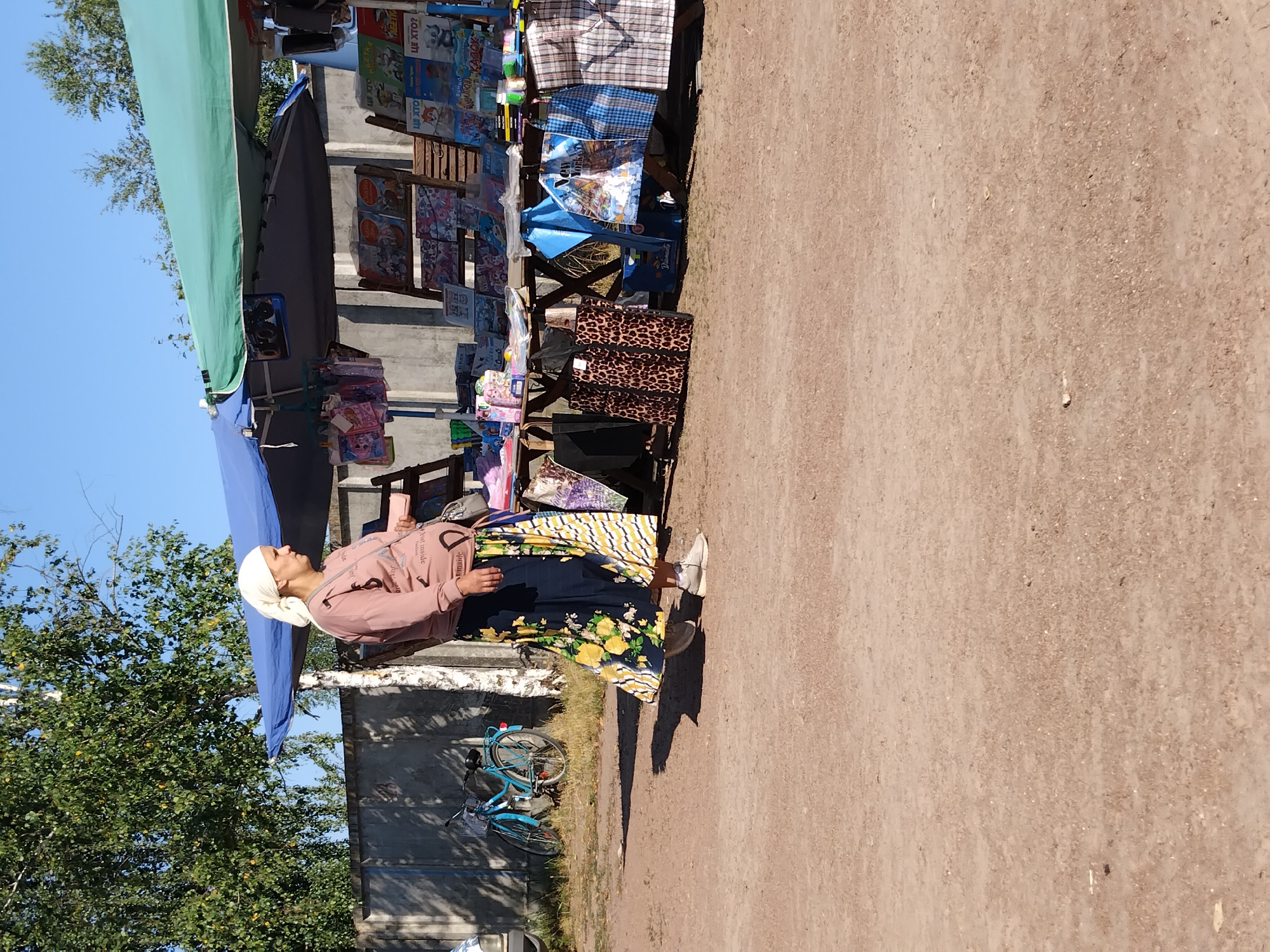 In Irshansk, Ukraine, a woman wearing a headscarf and a long, patterned skirt stands near a market stall. The stall, covered by blue and green awnings, displays various items, including books and bags. To the left of the stall, a light blue bicycle is parked. The ground is covered in dirt and gravel, and a concrete wall and trees are visible in the background, under a clear blue sky.