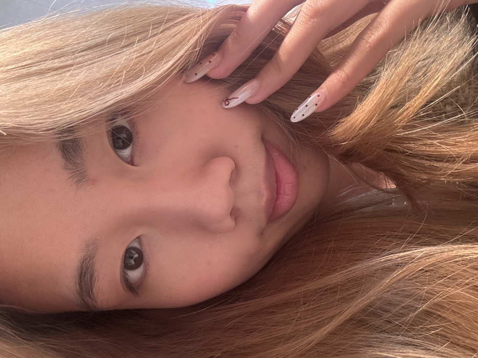 A close-up portrait depicts an East Asian individual, appearing to be a young female, with light brown, wavy hair partially framing her face. She has dark brown eyes and her lips form a subtle, closed-mouth smile. A small red blemish is visible on her forehead. Her right hand is raised, with manicured fingers resting near her right cheek and hair. Her fingernails are long and almond-shaped, featuring a clear or milky white base with dark red designs: the index finger displays a thin, irregular line, while the middle and ring fingers are adorned with small polka dots. The background is uniformly blurred and indistinct, offering no discernible environmental or architectural features. No visual information within the image identifies the location as Shapingba District, China, or any specific geographic area.