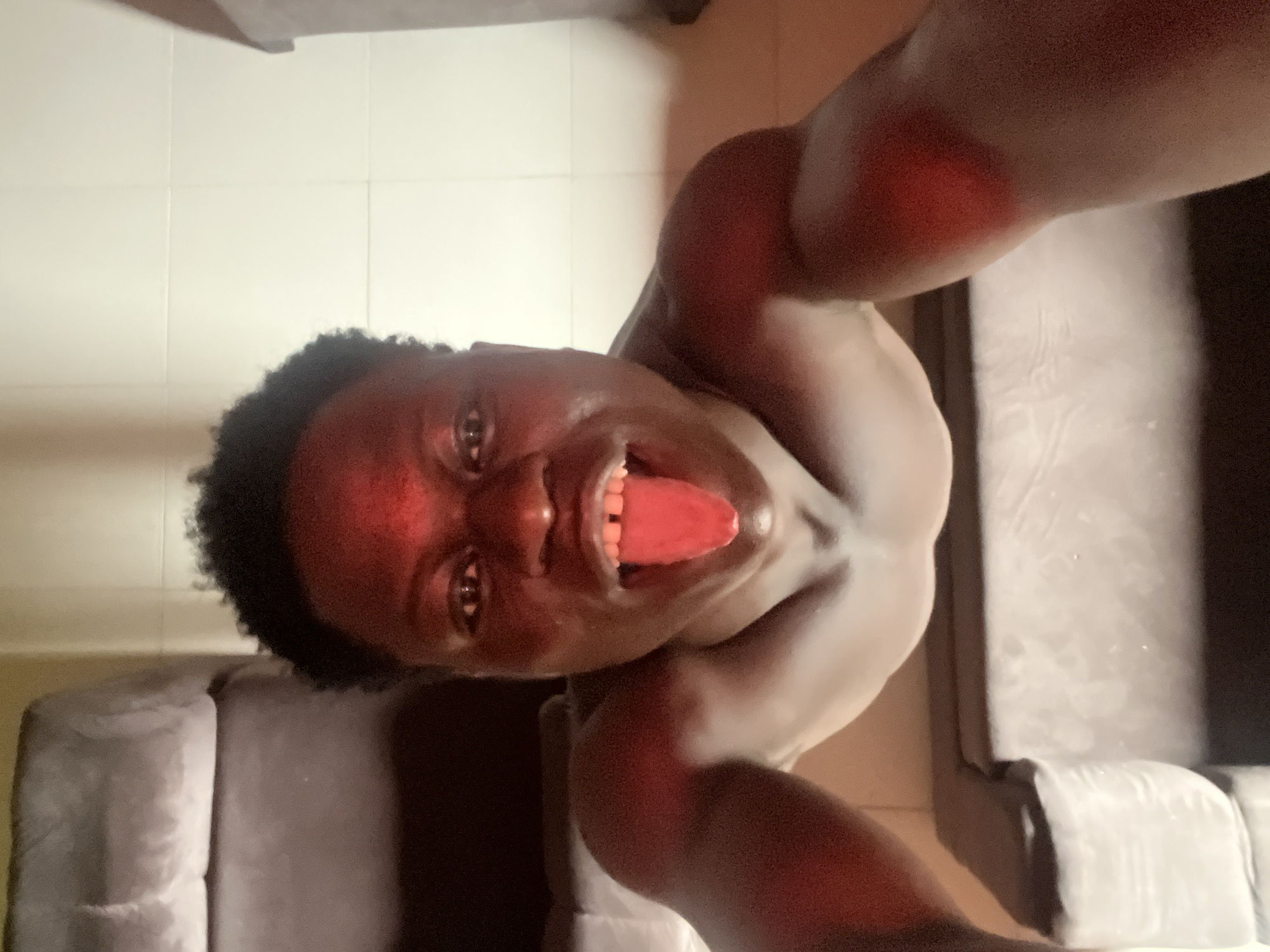 A dark-skinned person is positioned in an upside-down view, with their head near the top-left corner of the image and their bare torso angled diagonally across the center of the frame. The person has short, dark curly hair, and their mouth is open with the tongue extended. Part of a gray couch is visible in the lower-left corner, while another gray couch runs along the right side of the image. The background features light-colored tiled flooring. The location appears to be indoors. The city of Mosafejaw, Nigeria is not deducible from the provided image.